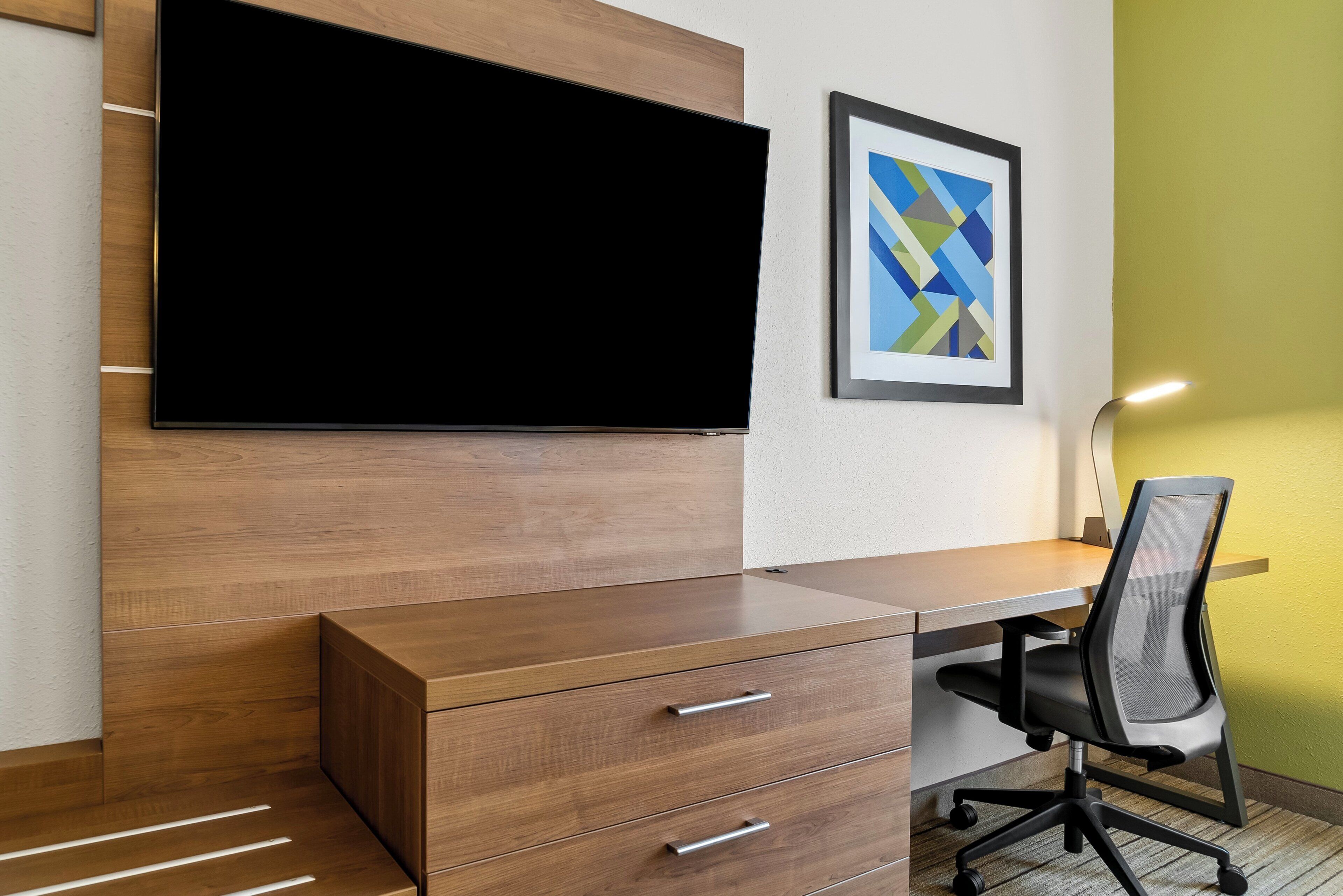 A modern office space with a large flat-screen TV mounted on a wooden wall, a desk with drawers, a black office chair, and a framed abstract painting on the wall.