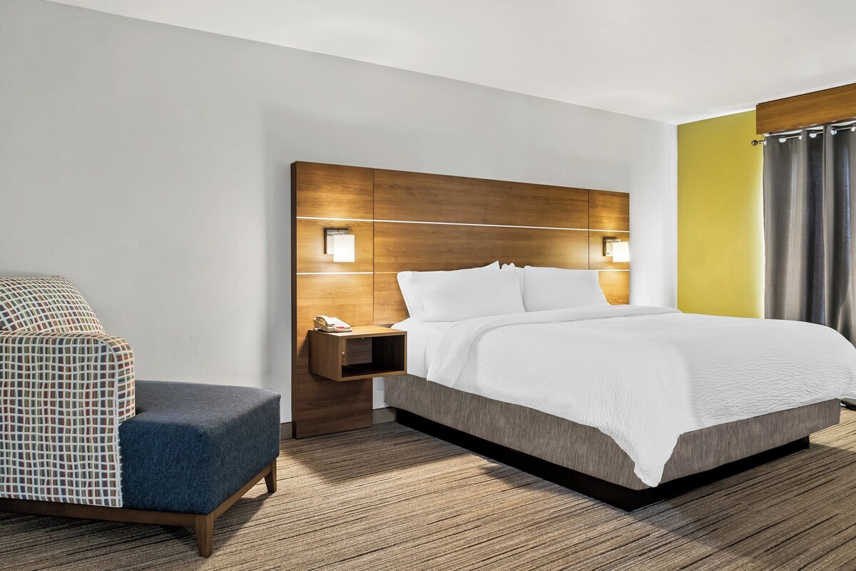 A modern hotel room with a large bed, a nightstand with a lamp, a chair, and a wooden headboard.