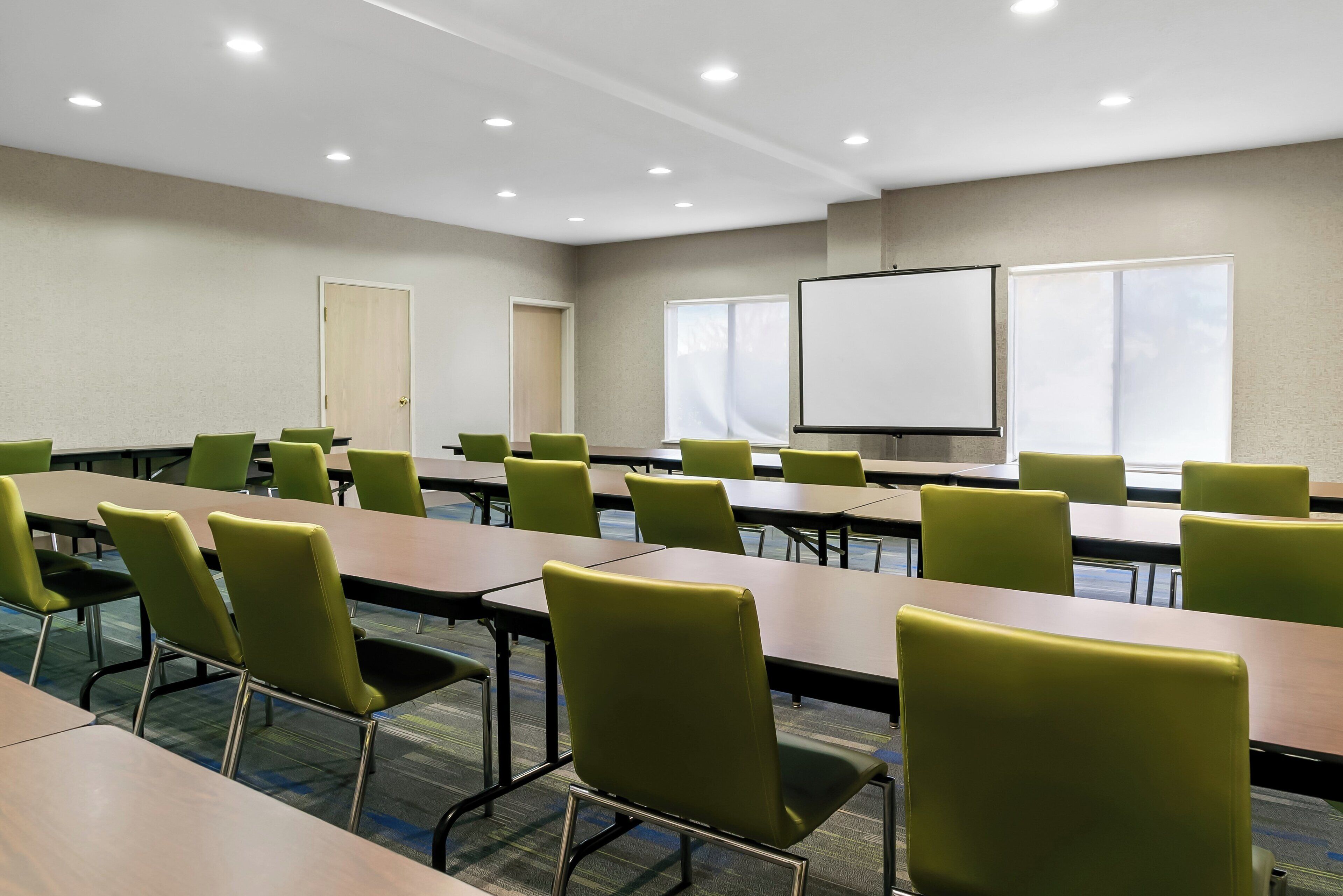 Une salle de conférence avec des chaises et des tables vertes, un tableau blanc et des fenêtres équipées de stores.