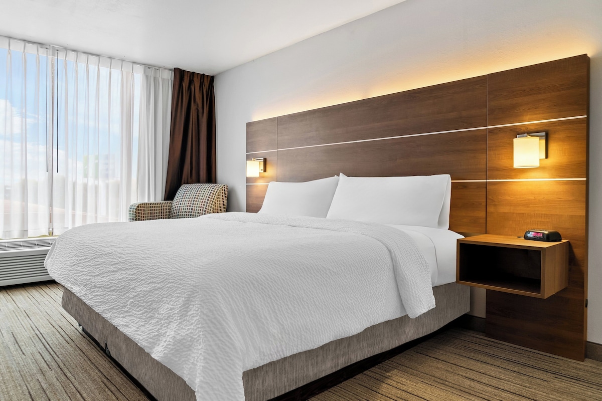Habitación de hotel con una cama grande, una mesita de noche con un reloj, una silla y una ventana con cortinas.