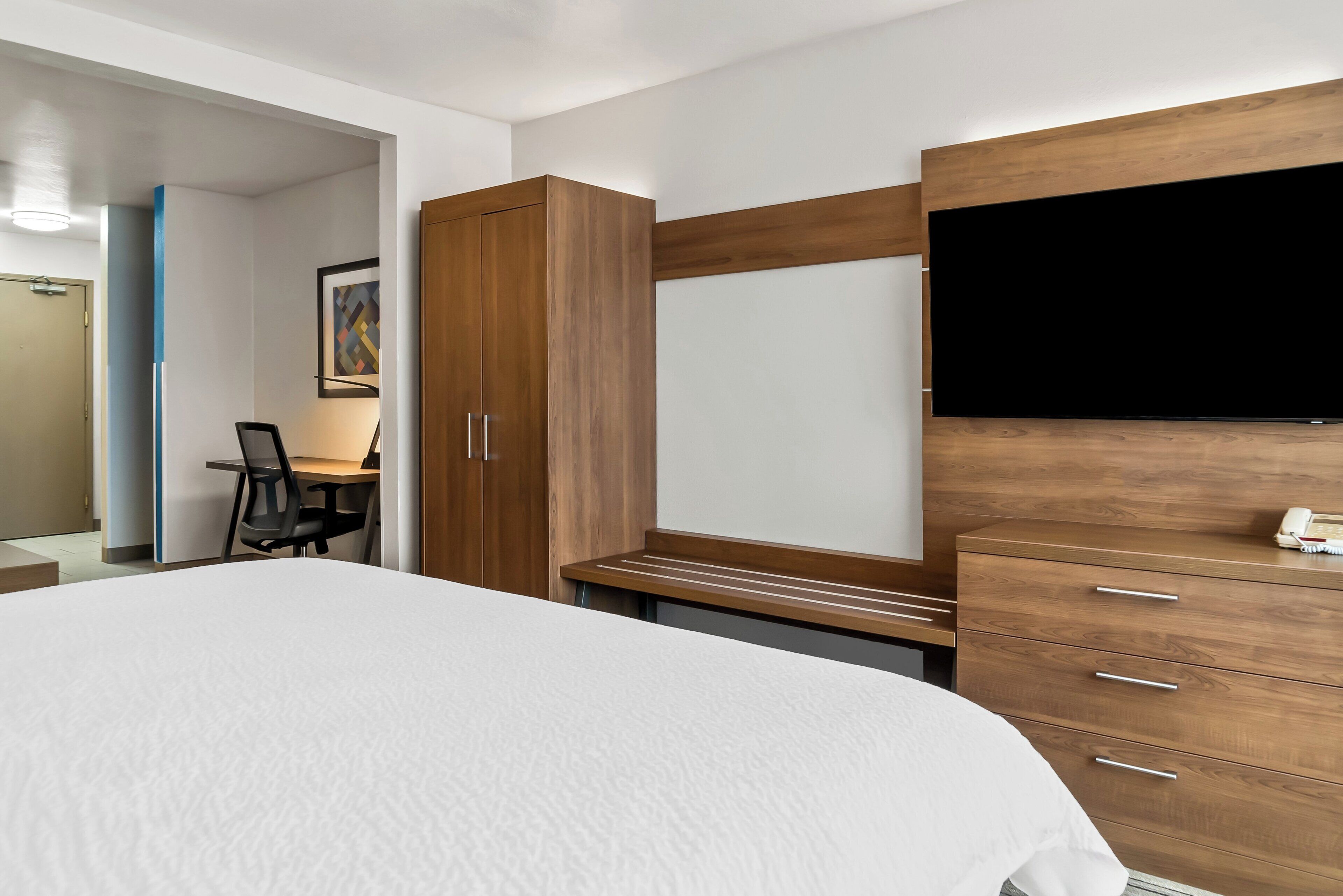A hotel room with a large bed, wooden wardrobe, desk, chair, and a flat-screen TV mounted on the wall.