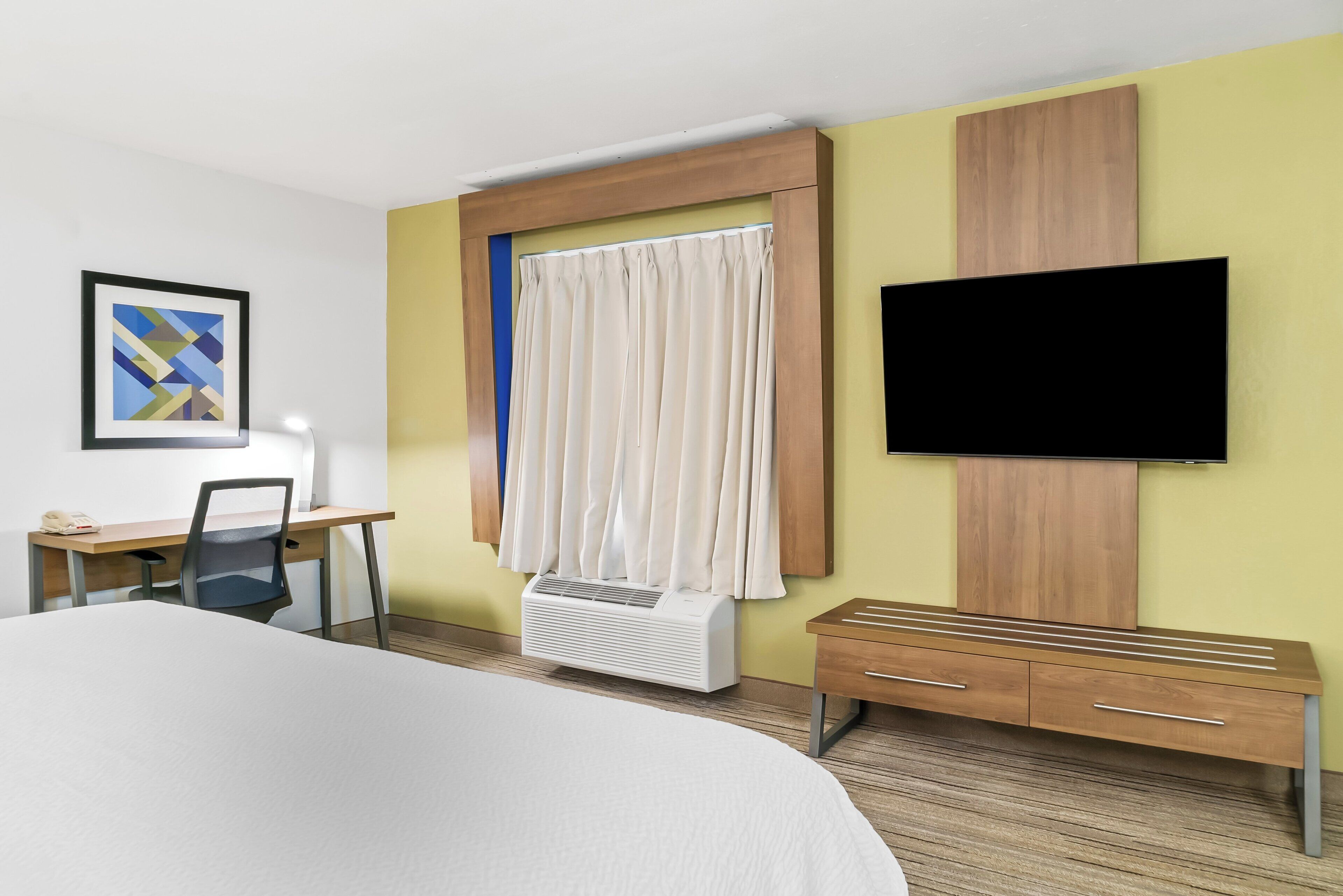 A hotel room with a bed, desk, chair, TV, air conditioning unit, and a wooden bench.