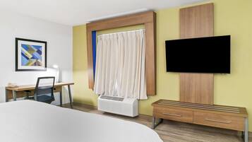 A hotel room with a bed, desk, chair, TV, air conditioning unit, and a wooden bench.