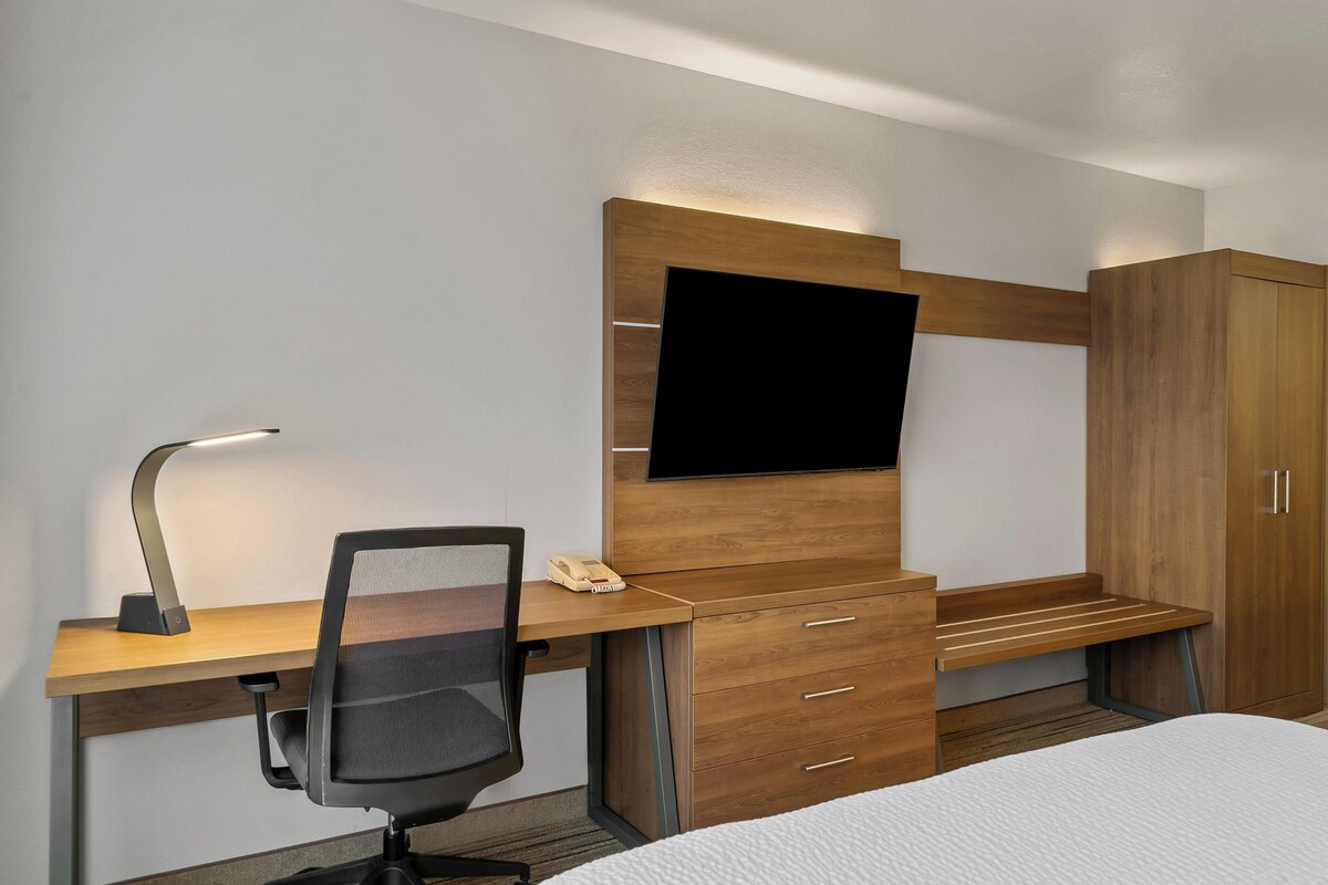 A modern hotel room with a wooden desk, a black office chair, a flat-screen TV mounted on the wall, and a wardrobe.