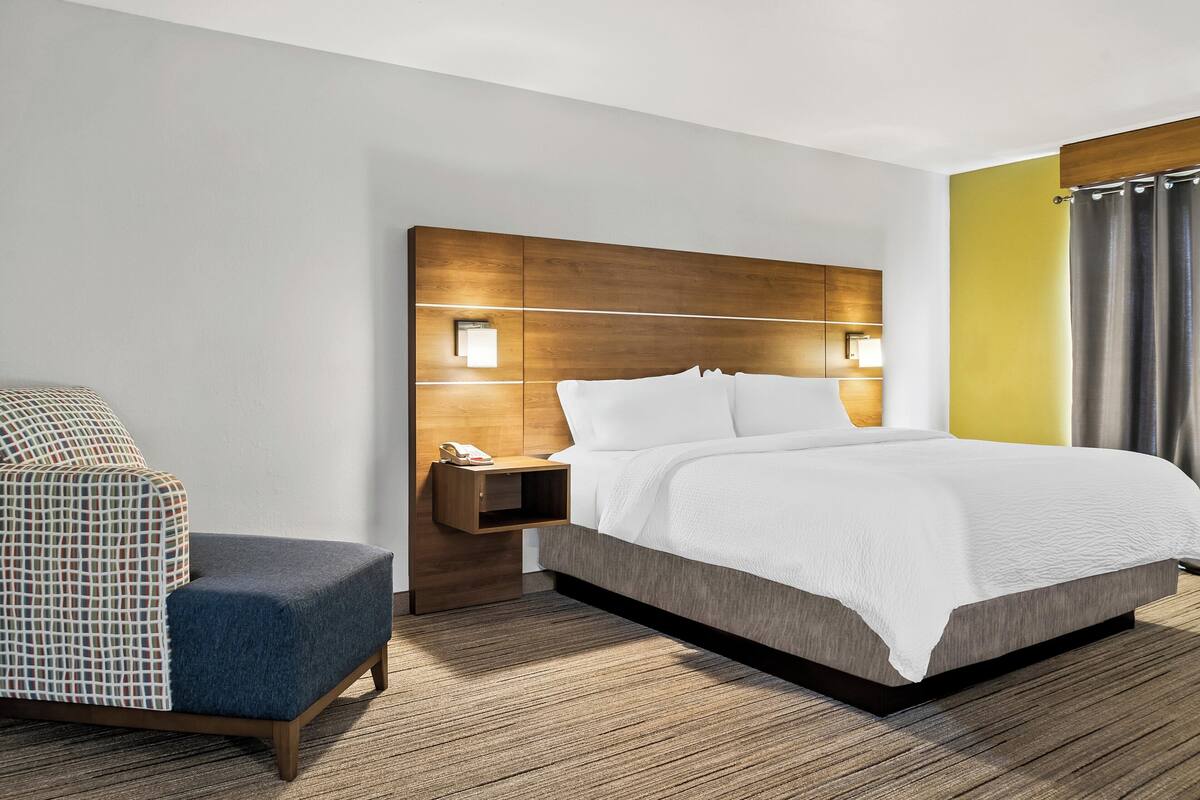 A modern hotel room with a large bed, a nightstand with a lamp, a chair, and a wooden headboard.