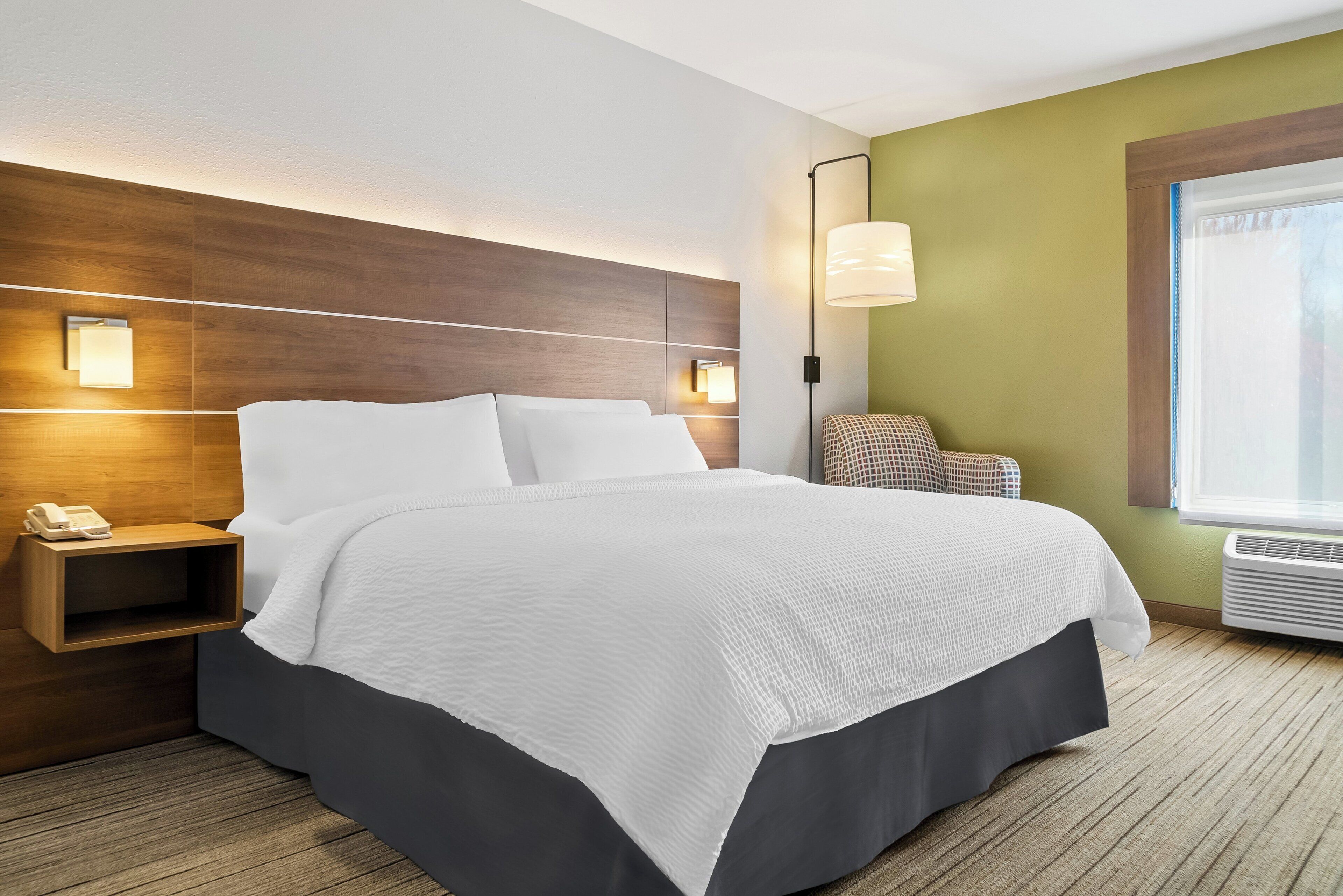A hotel room with a large bed, bedside lamps, a small table with a phone, and a window with blinds.