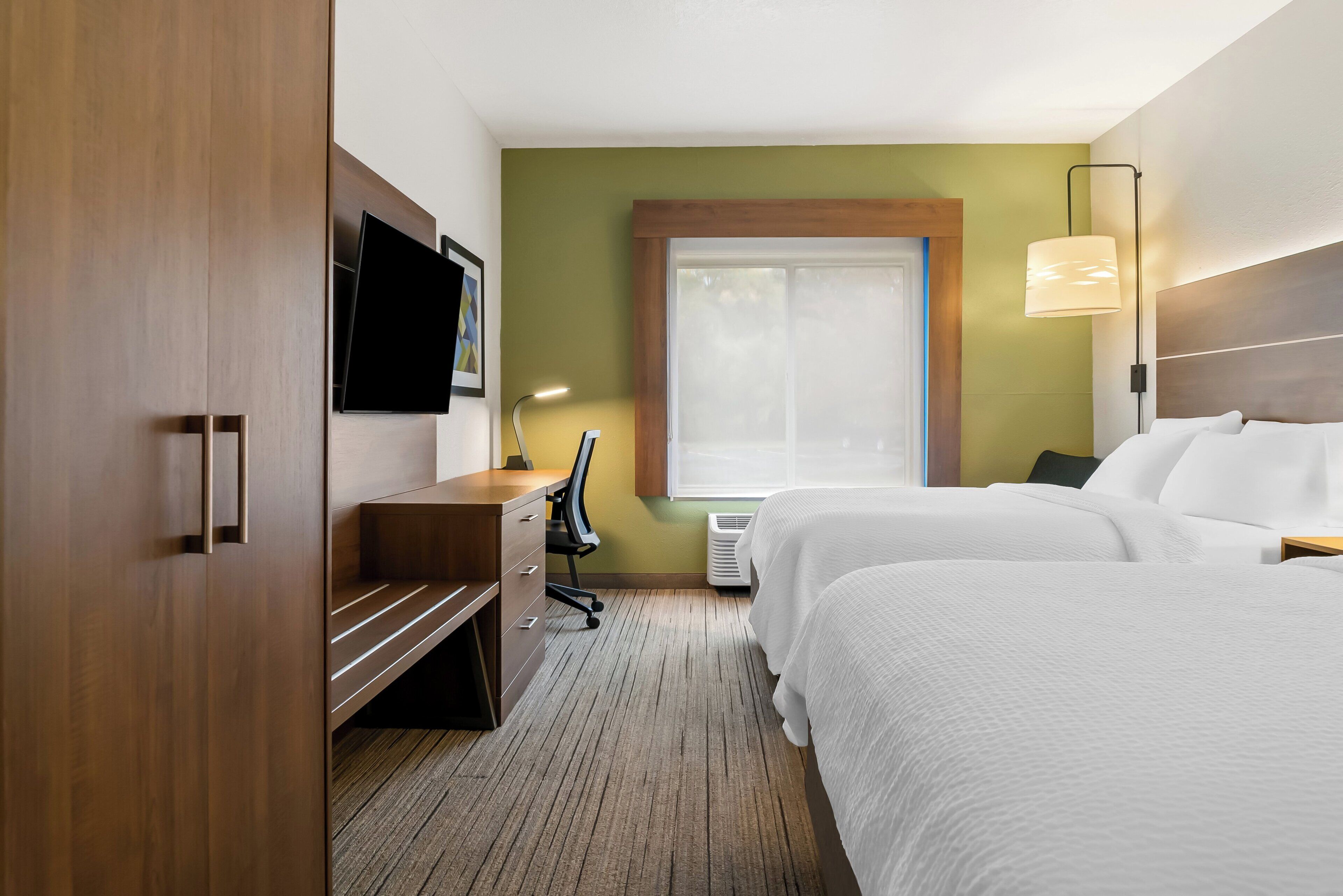 A hotel room with a wooden wardrobe, a desk with a chair, a flat-screen TV, two beds, and a window with a view.