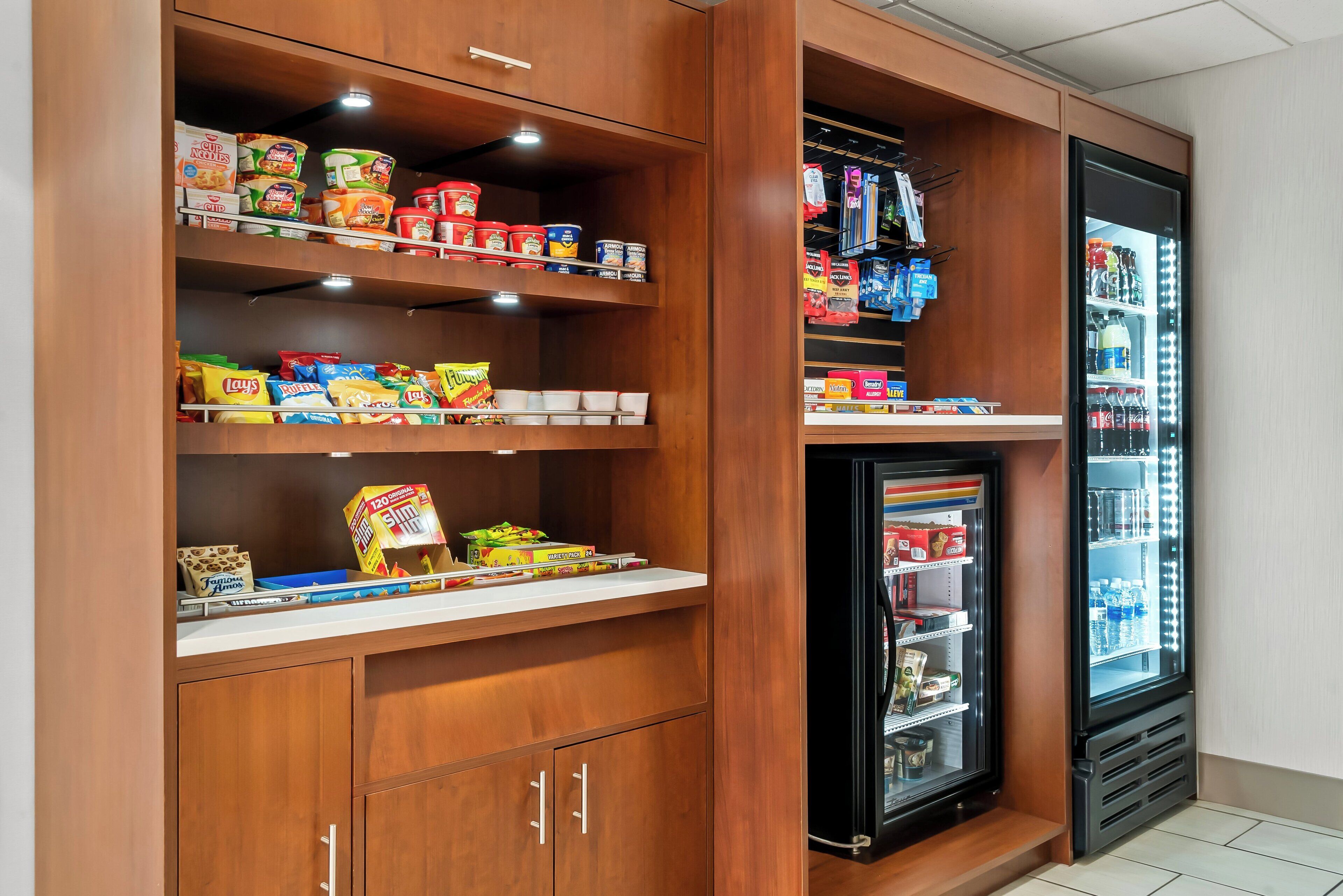 A kitchenette with a refrigerator stocked with beverages and a shelf with canned goods and snacks.