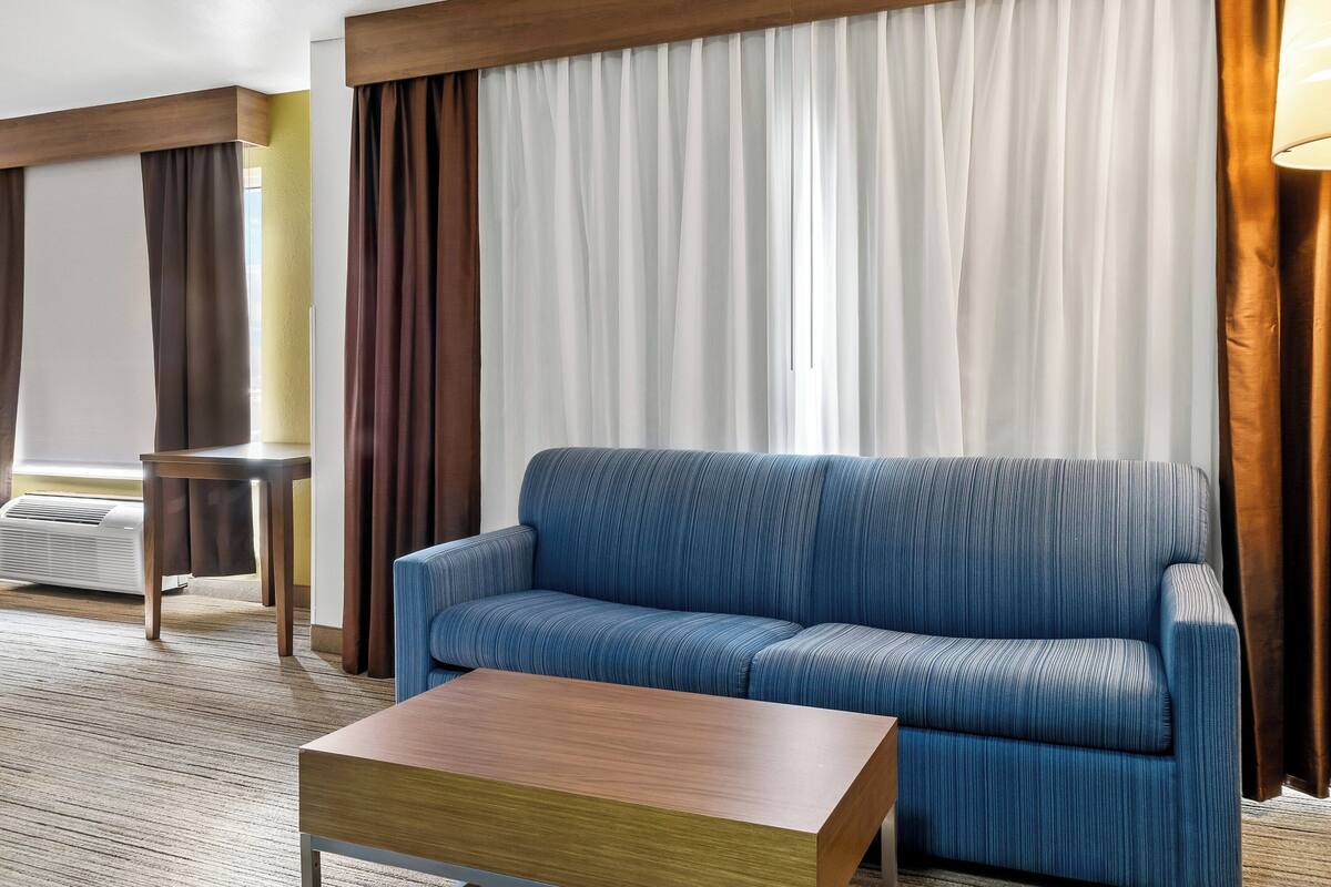 A modern hotel room with a blue sofa, a wooden coffee table, and a large window with curtains.