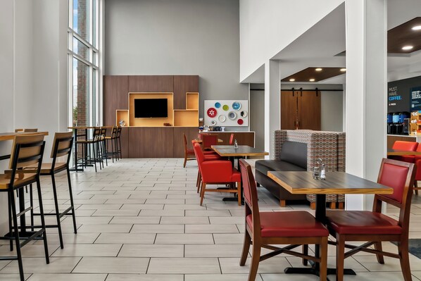 Un café moderne avec des tables en bois et des chaises rouges, un téléviseur à écran plat et un mur décoré d’assiettes colorées.