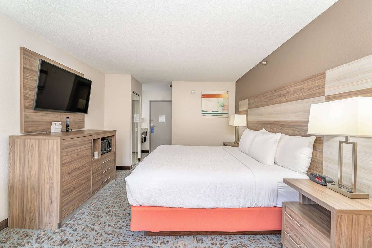 A hotel room with a large bed, a TV mounted on a wooden panel, a desk with a lamp, and a small table with a clock.