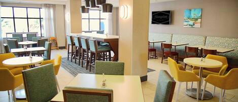 A hotel dining area with multiple tables and chairs, a bar, and a television on the wall.