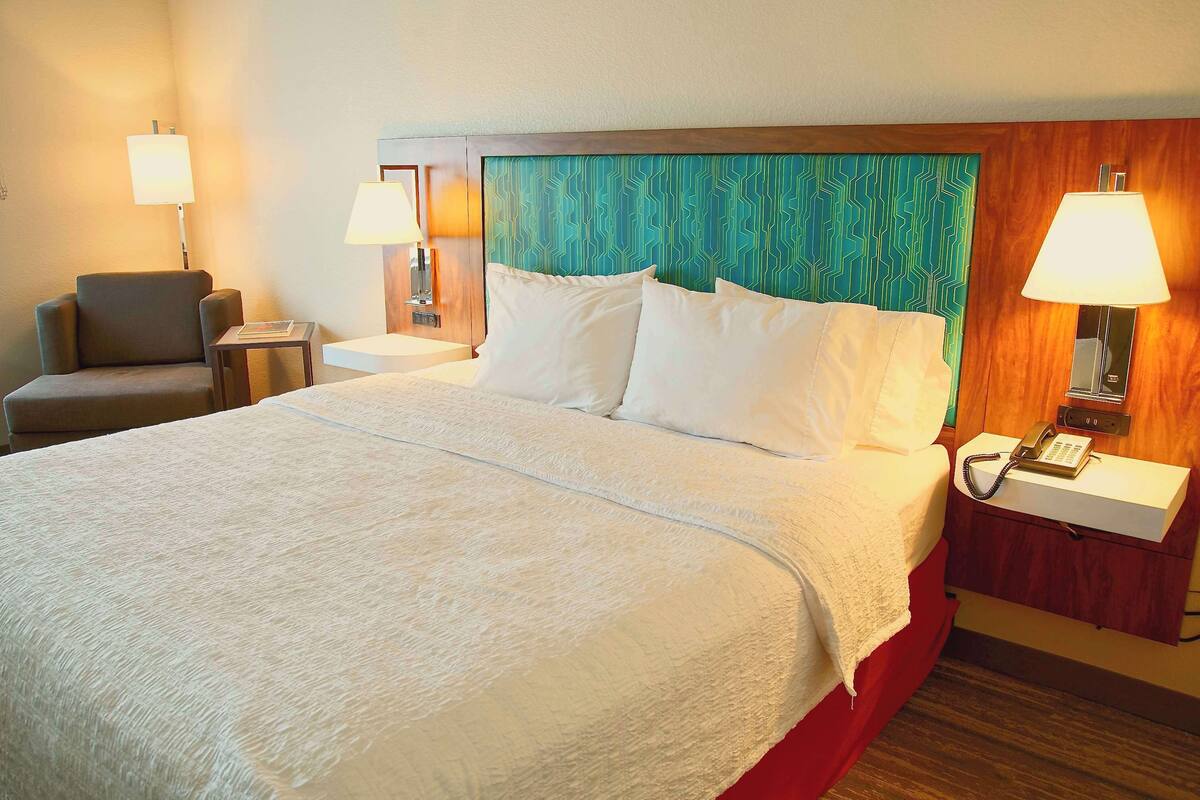 A hotel room with a large bed, two bedside tables with lamps, a chair, and a wall with a patterned headboard.