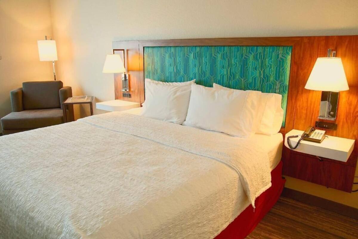 A hotel room with a large bed, two bedside tables with lamps, a chair, and a wooden headboard.
