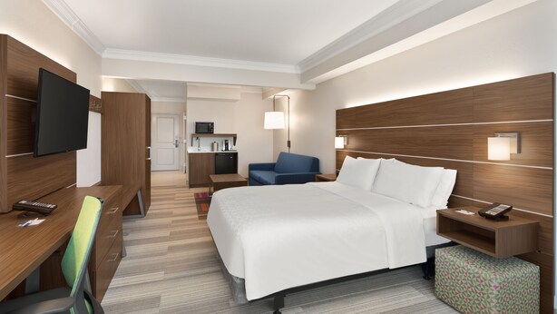 A hotel room with a bed, a desk with a chair, a TV, a bedside table, a lamp, and a small kitchenette in the background. - Holiday Inn Express & Suites Byron by IHG (Byron)