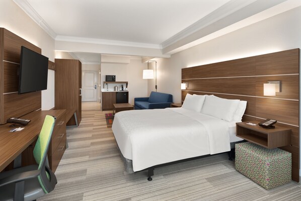 Hotelkamer met een bed, een bureau met stoel, een televisie, een nachtkastje, een lamp en een kleine kitchenette op de achtergrond.