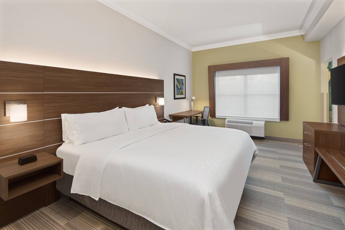 A hotel room with a large bed, bedside tables, a desk, a chair, a TV, and a window with blinds.