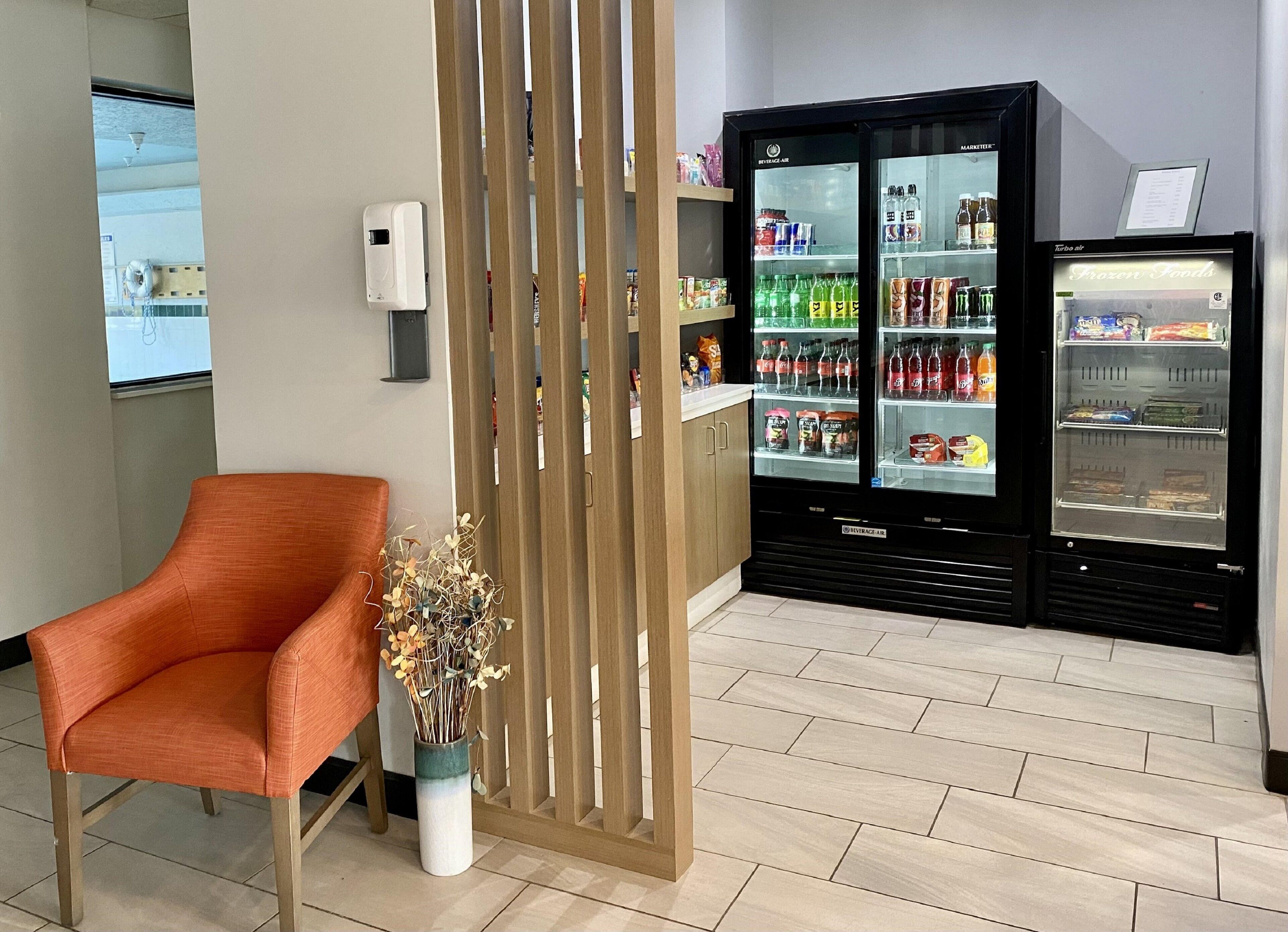 A seating area with a modern orange chair, a decorative plant, and a beverage vending machine with a freezer section.