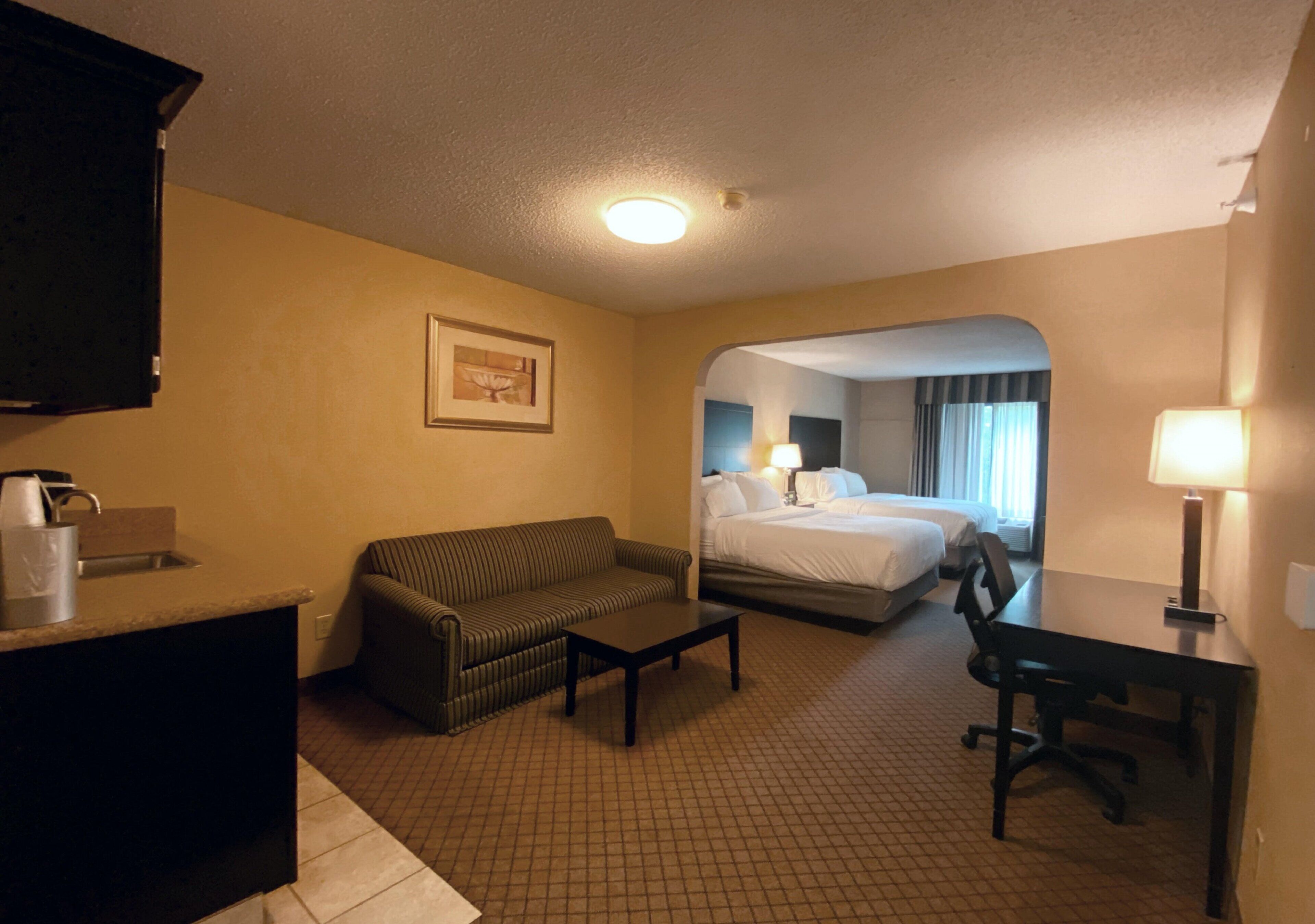 A hotel room with a sofa, a small table, a desk, and a bed. There is a picture on the wall and a window with curtains.