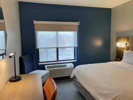 Habitación de hotel con cama, escritorio, silla, ventana con vista a un estacionamiento y una lámpara de pared.