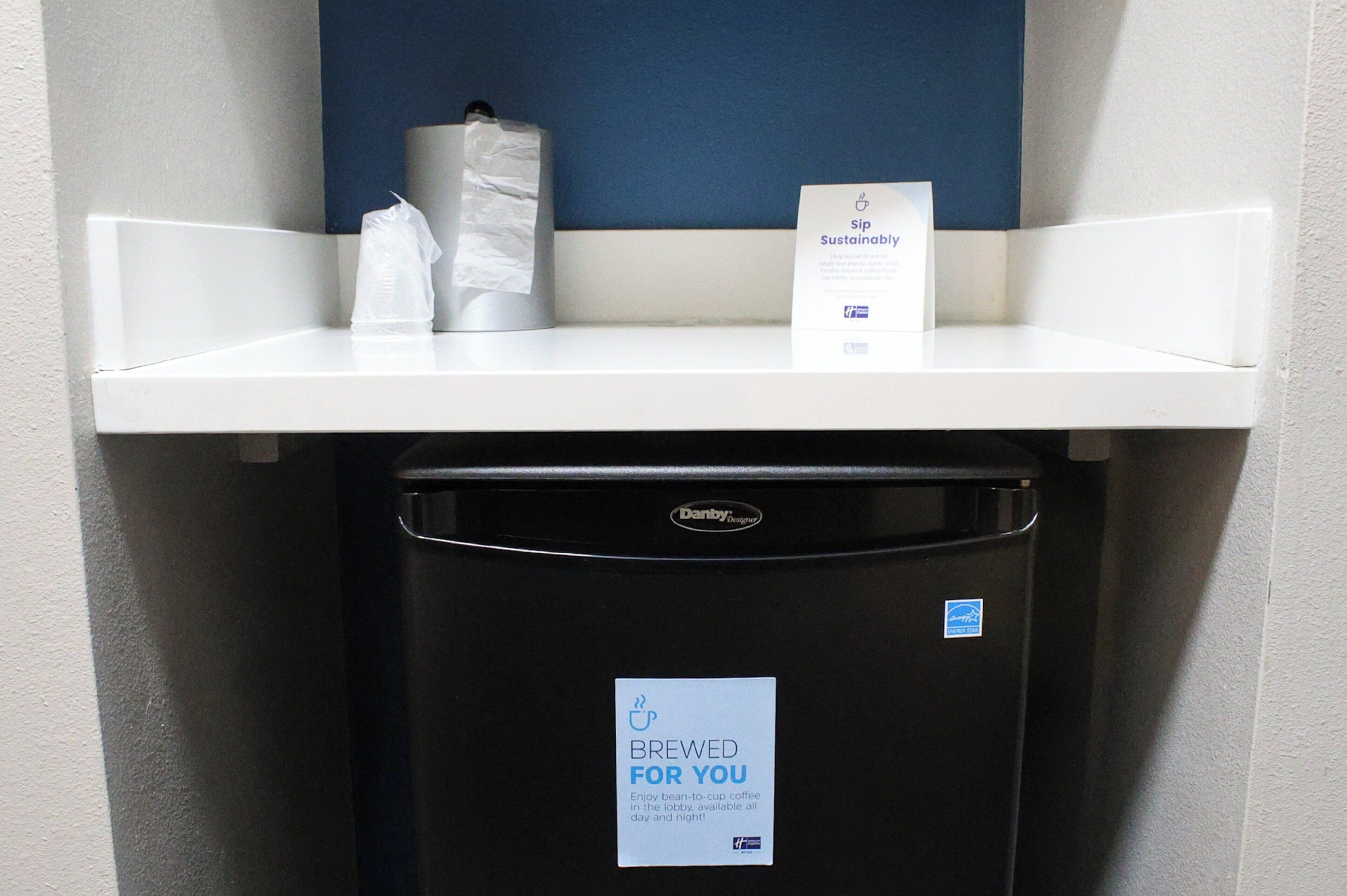 Une machine à café avec une étiquette indiquant que les grains de café proviennent de sources durables, une poubelle et un distributeur d’essuie-tout.