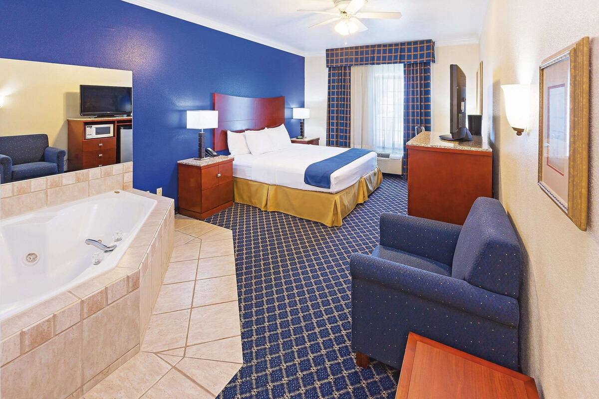 A hotel room with a large bed, a sitting area, a TV, a desk, and a bathroom with a bathtub.