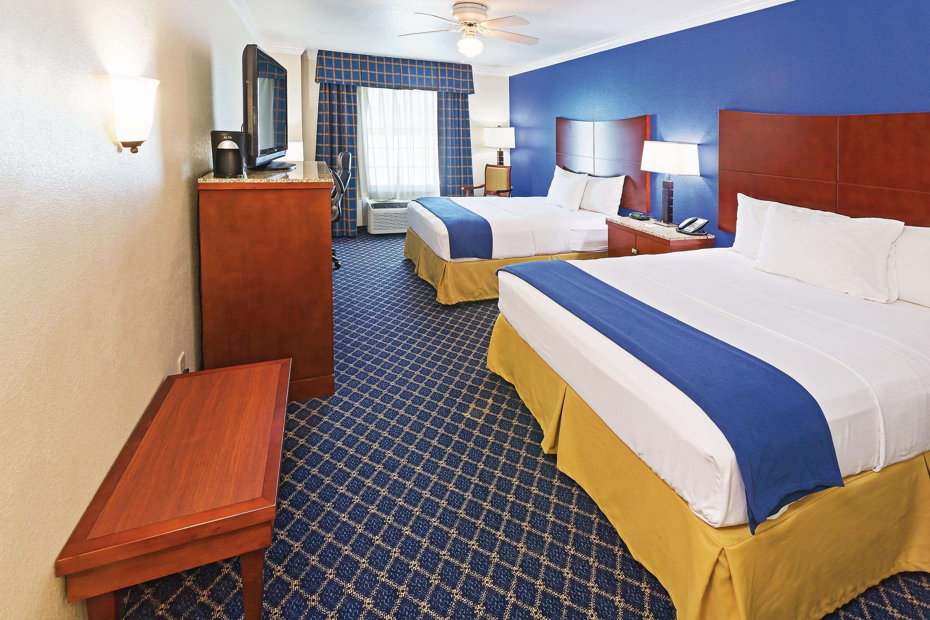 A hotel room with two beds, a desk, a chair, a TV, a lamp, and a ceiling fan.