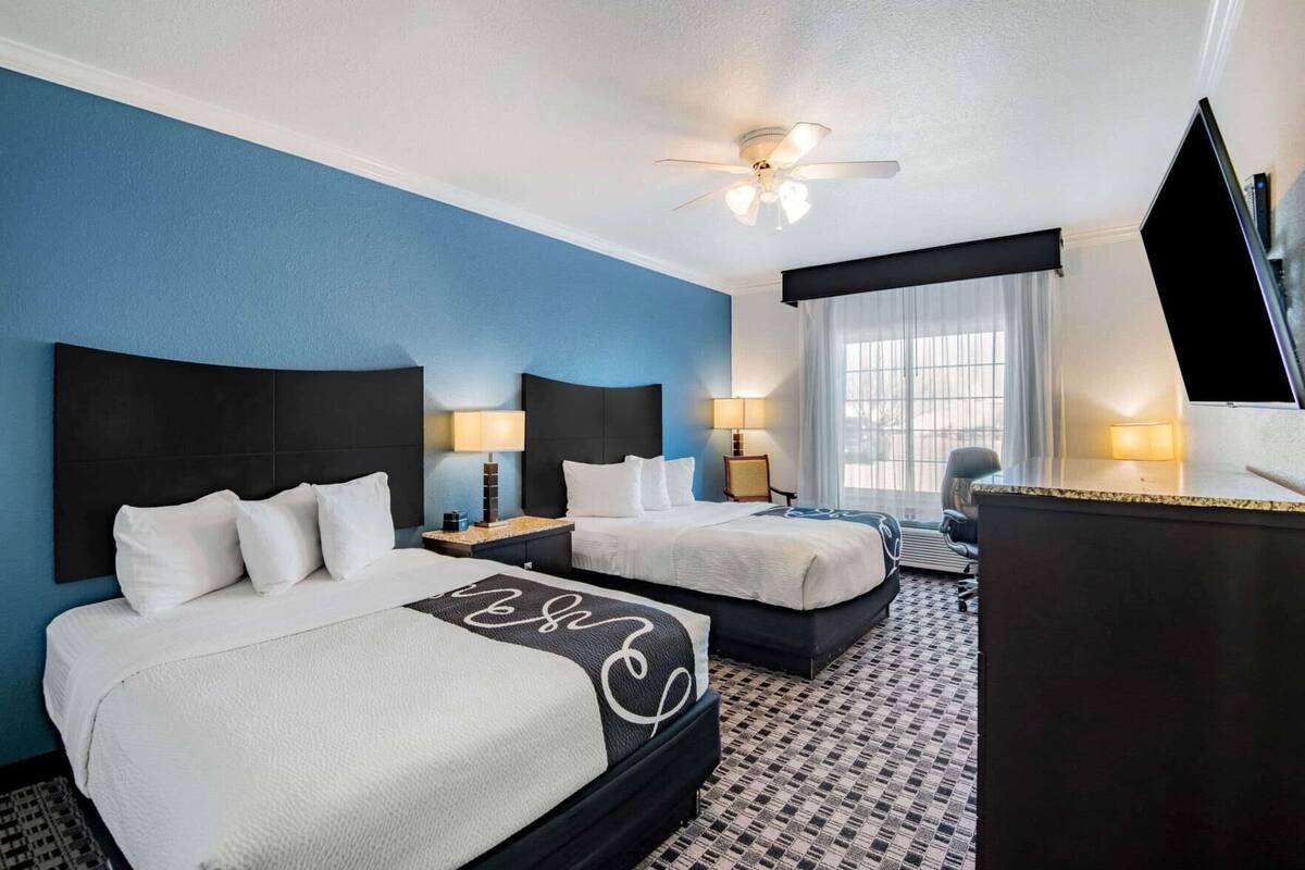 A hotel room with two beds, a desk, a chair, a TV, and a ceiling fan.