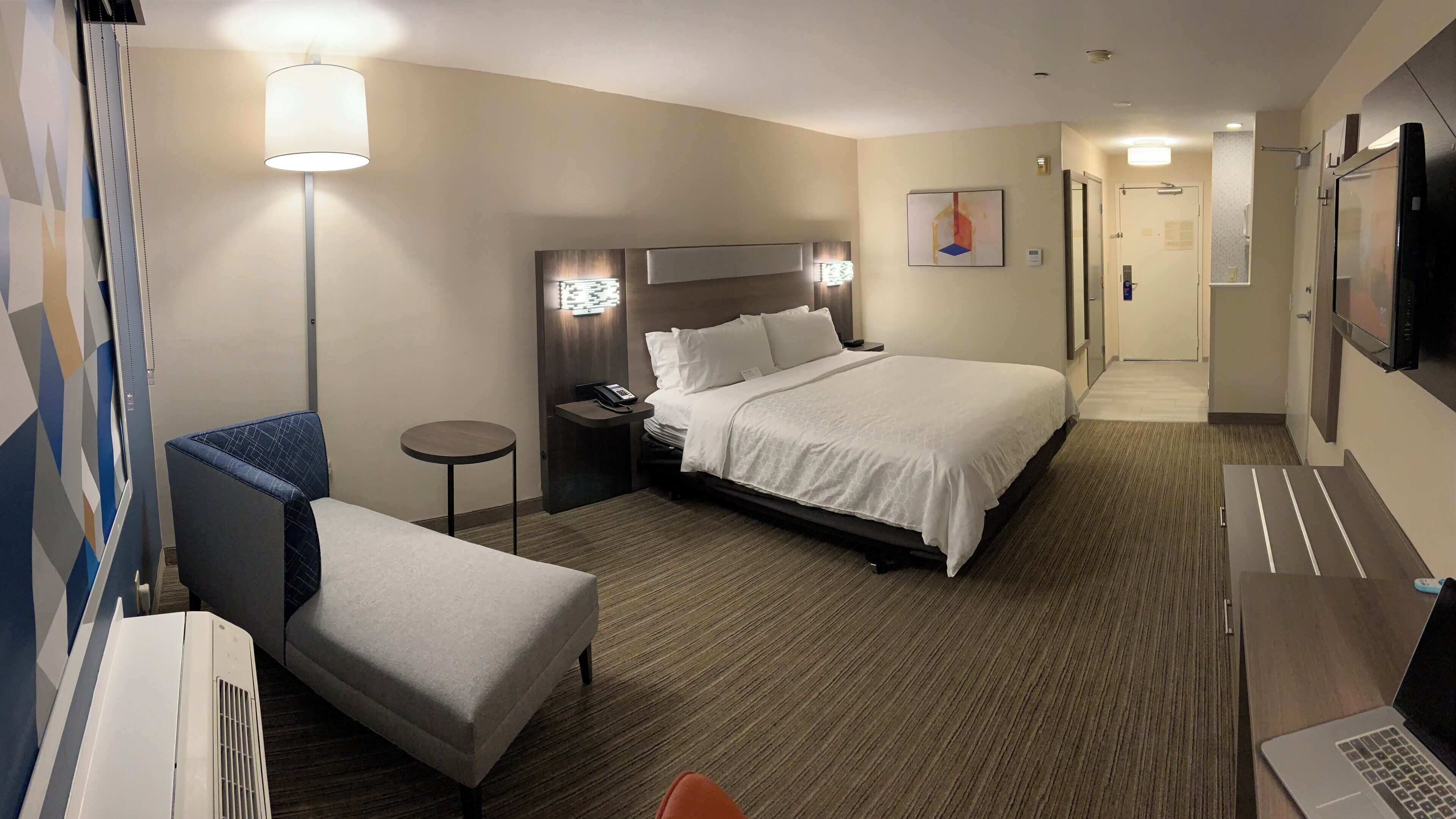A hotel room with a bed, a desk, a chair, a lamp, a TV, and a small table.