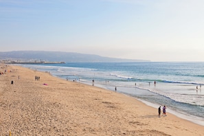 Platja ampla i sorrenca amb ones suaus, un moll a la distància i un cel clar.