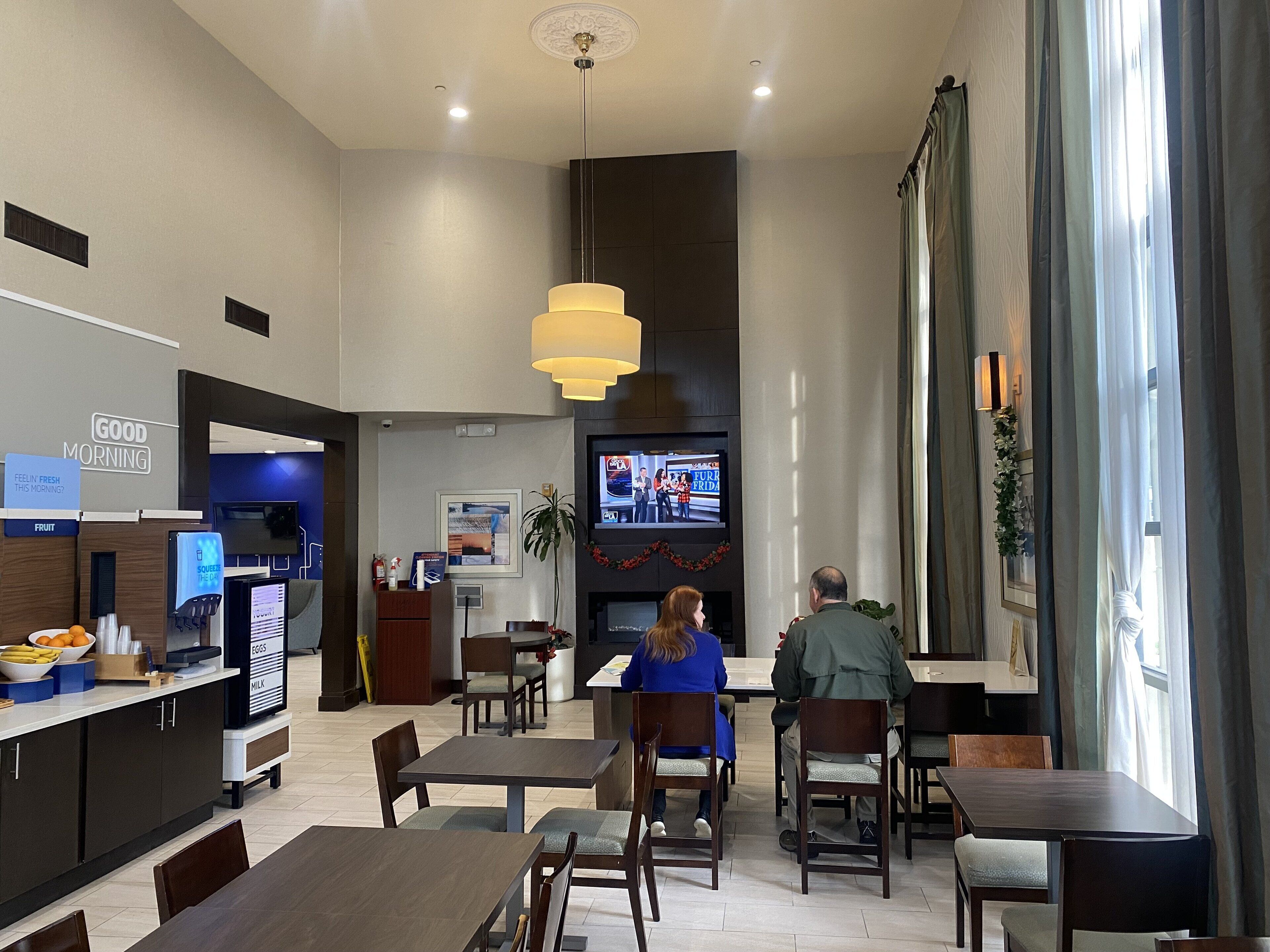 Een modern restaurant met een televisie waarop een nieuwsprogramma wordt vertoond, een fruitbar en een zithoek met tafels en stoelen.