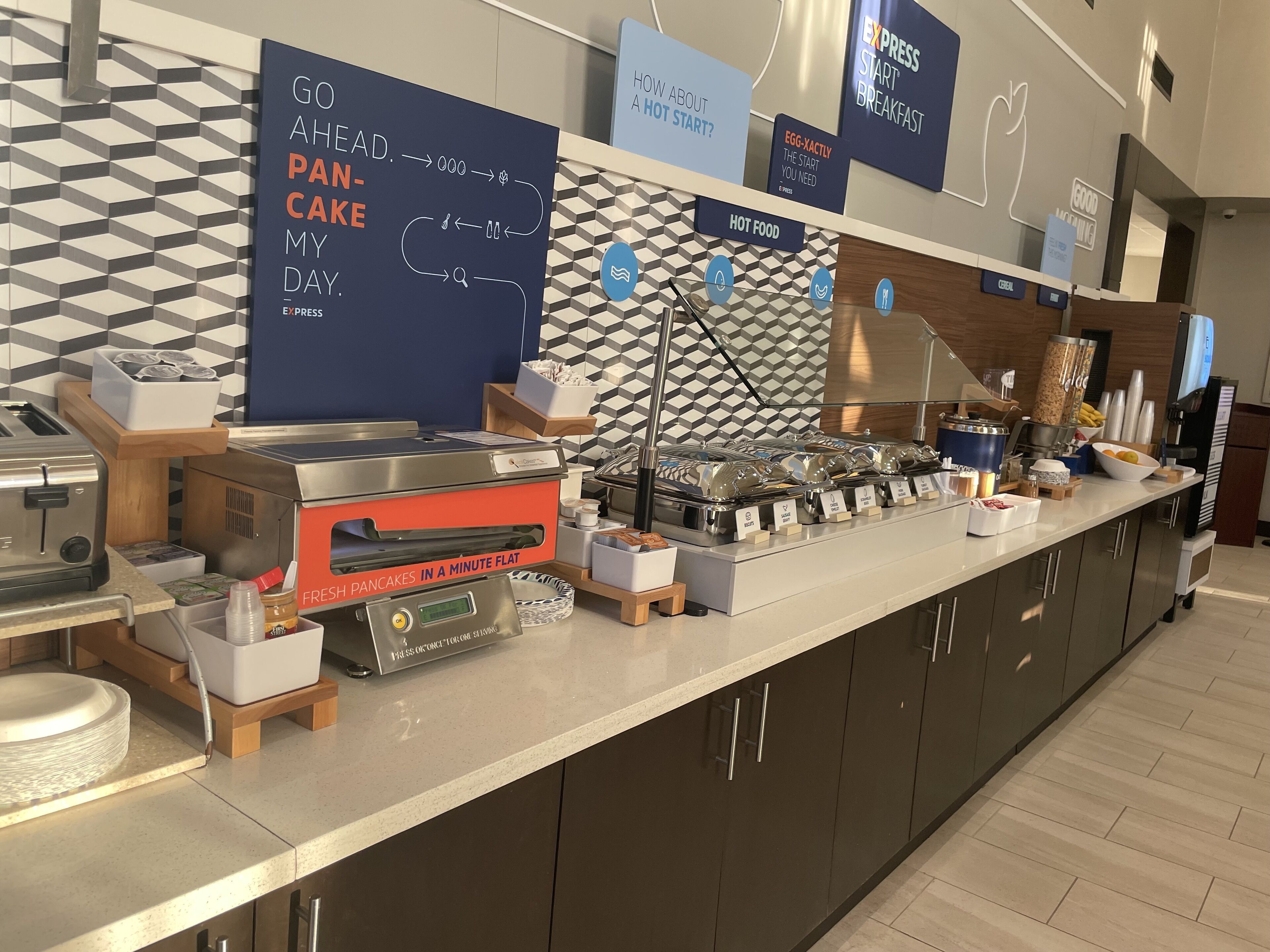 Zona de desayuno tipo bufé con plancha para panqueques, estación de café y varios recipientes de comida.