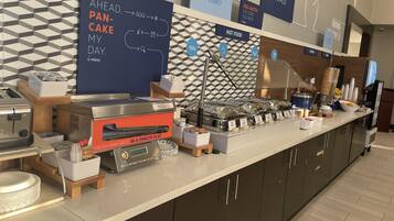 Zona de desayuno tipo bufé con plancha para panqueques, estación de café y varios recipientes de comida.