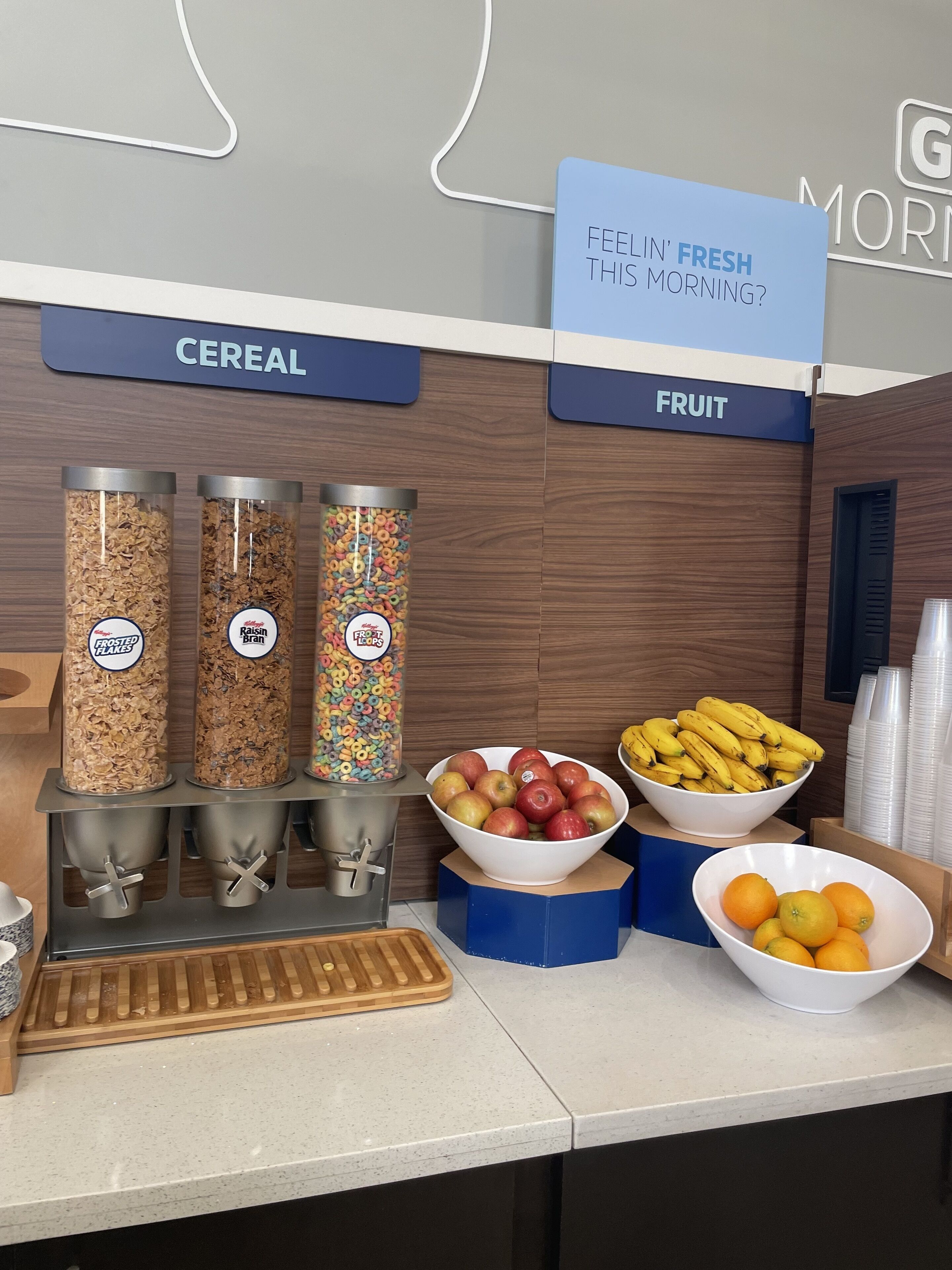 A breakfast station with labeled dispensers for cereal and fruit, including apples, bananas, and oranges.