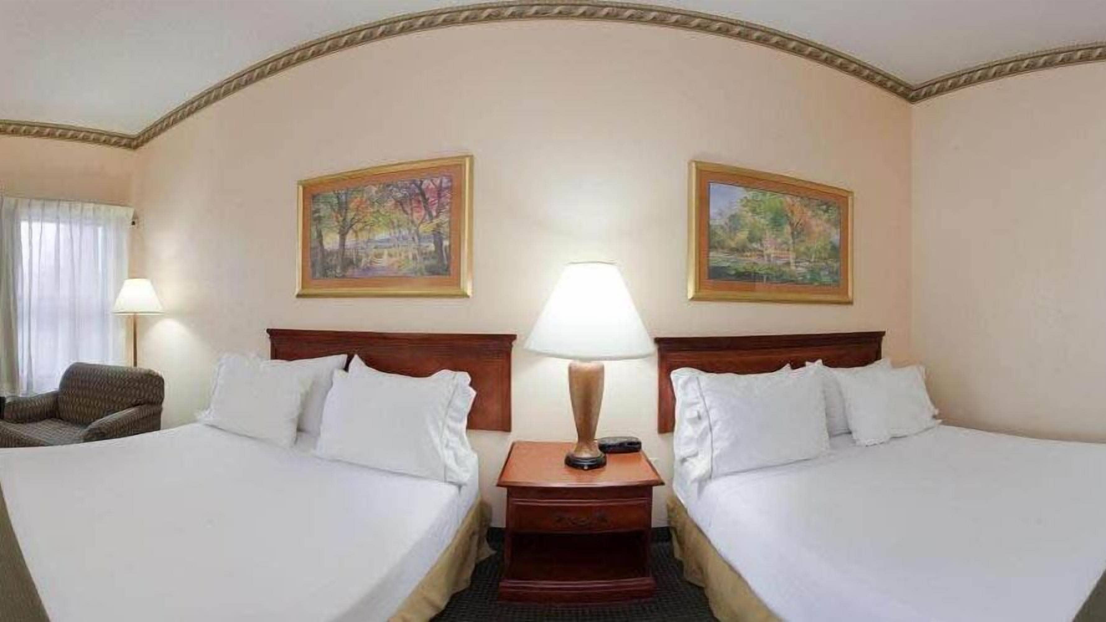 A hotel room with two beds, a nightstand with a lamp, a chair, and two paintings on the wall.