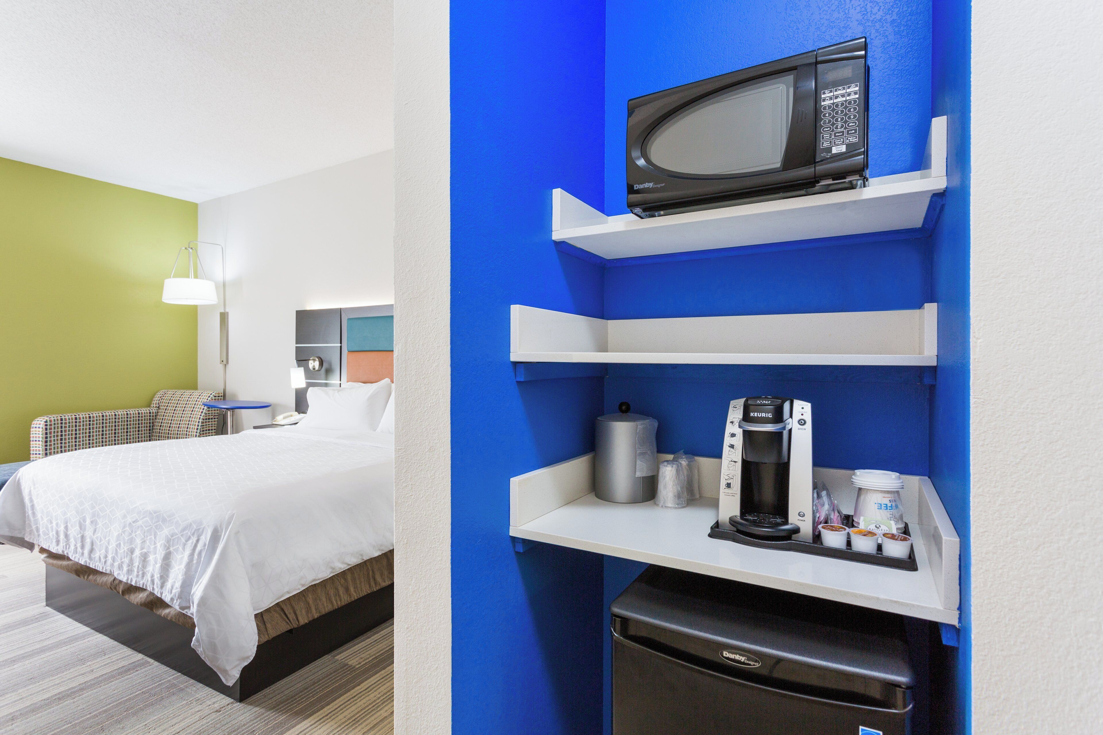 A hotel room with a bed, a desk, a microwave, a coffee maker, and a trash bin.