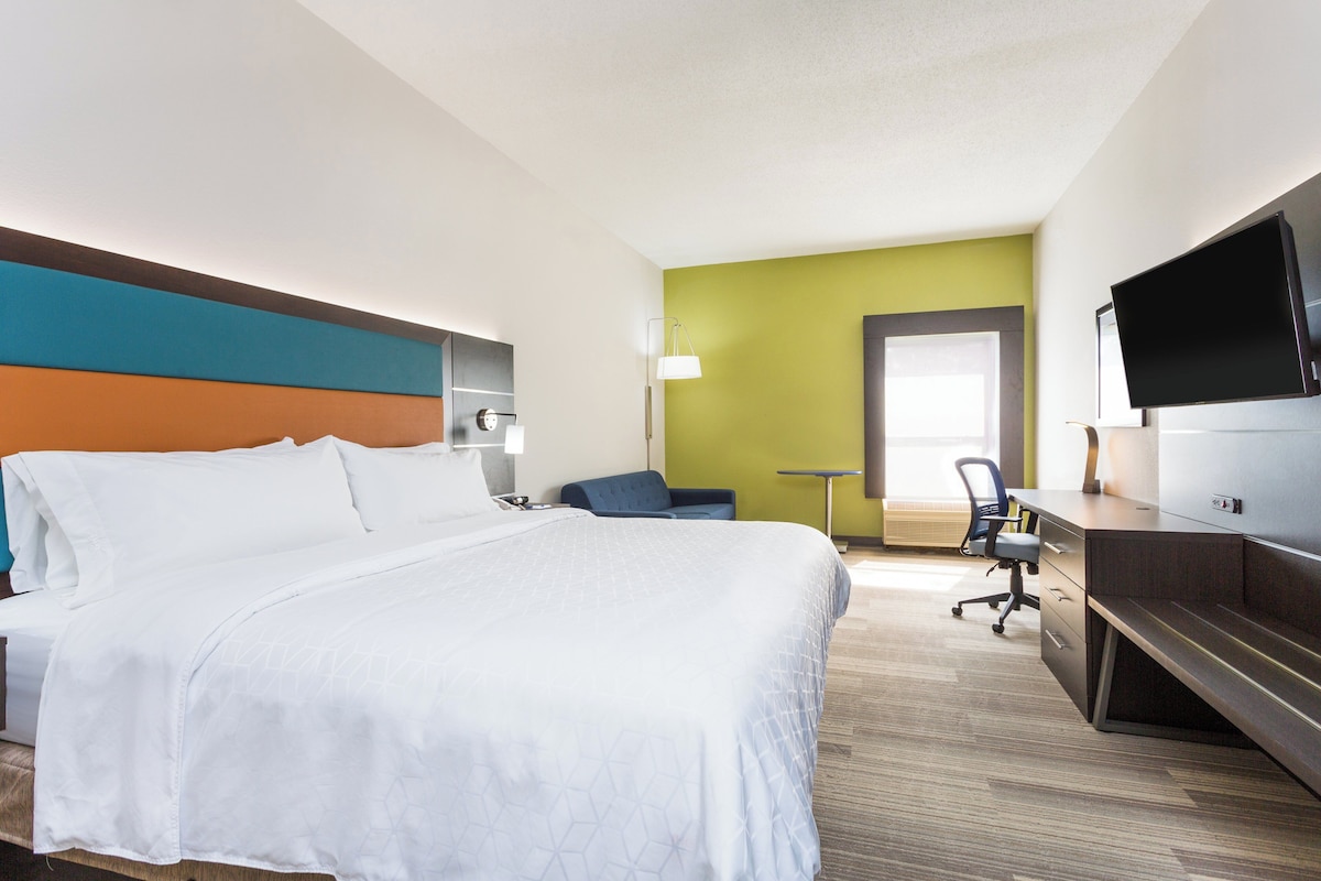 A hotel room with a large bed, a desk, a chair, a television, and a door leading to another room.