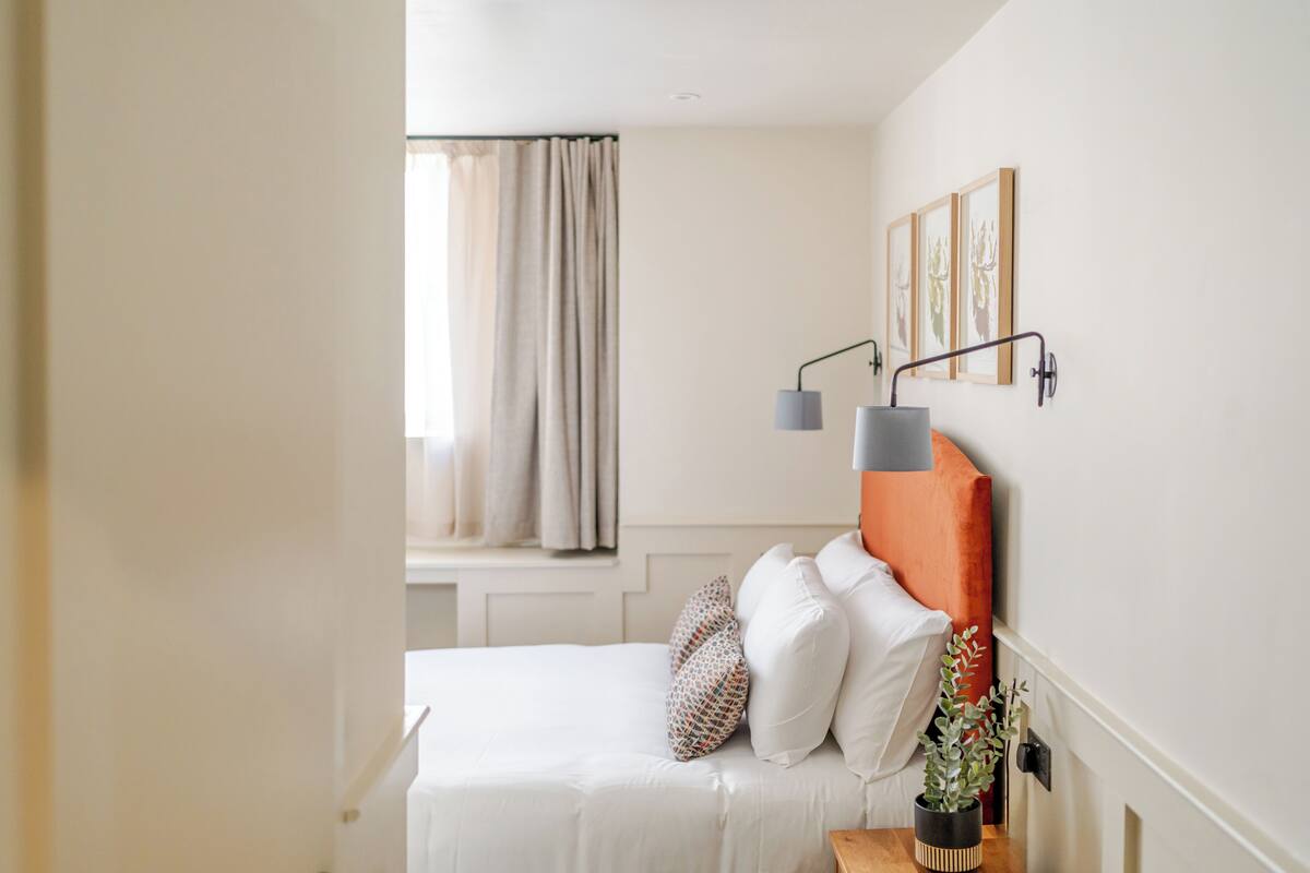 A bedroom with a bed, a bedside table, a lamp, a window with curtains, and wall-mounted artwork.