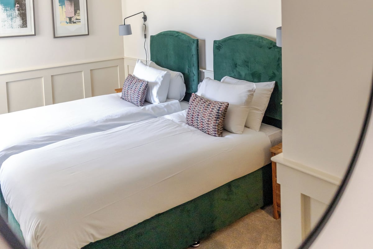 A bedroom with two beds, a green headboard, white bedding, a bedside table with a lamp, and two framed pictures on the wall.
