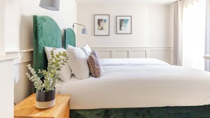 A neatly made bed with white linens, a green headboard, and a bedside table with a plant. There are two framed pictures on the wall above the bed.