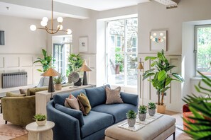 A modern living room with a blue sofa, green armchair, and a beige ottoman. There are several potted plants and a large window providing natural light. - Grove House Hotel (Edinburgh)