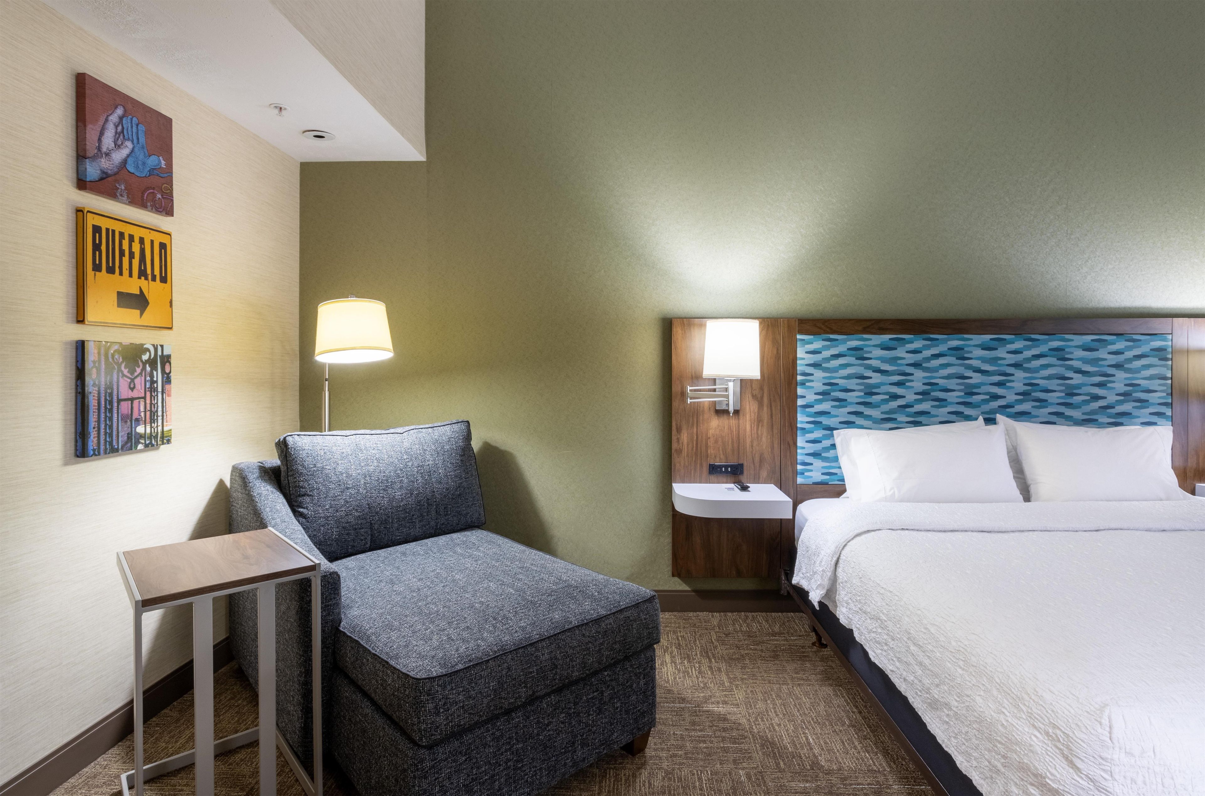 A hotel room with a bed, a nightstand, a lamp, a chair, and a small table.