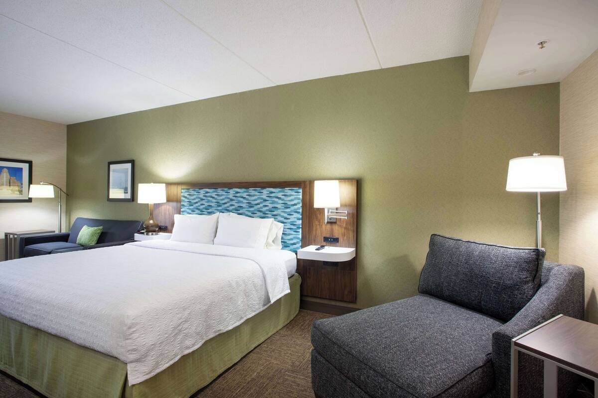 Une chambre d’hôtel avec un grand lit, un fauteuil gris, une table de chevet avec une lampe et une étagère fixée au mur.