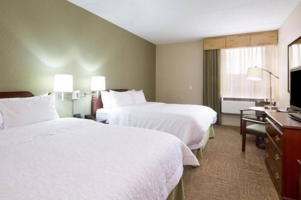 Une chambre d’hôtel avec deux lits, des lampes de chevet, un bureau avec une chaise, une télévision et une fenêtre avec des rideaux.