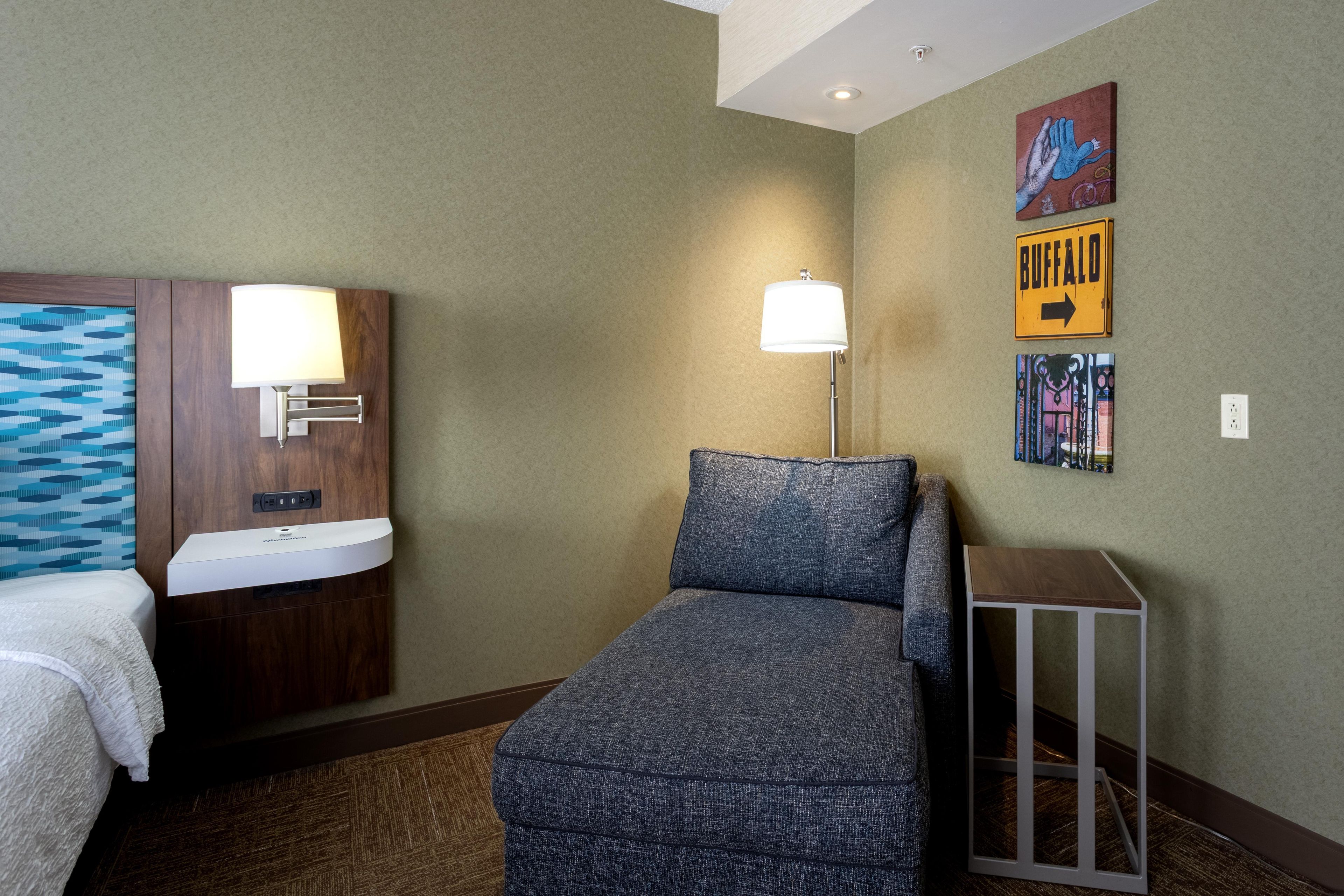Une chambre d’hôtel comprenant un lit, un petit canapé, une table de chevet, une lampe, une étagère fixée au mur et une œuvre d’art décorative.