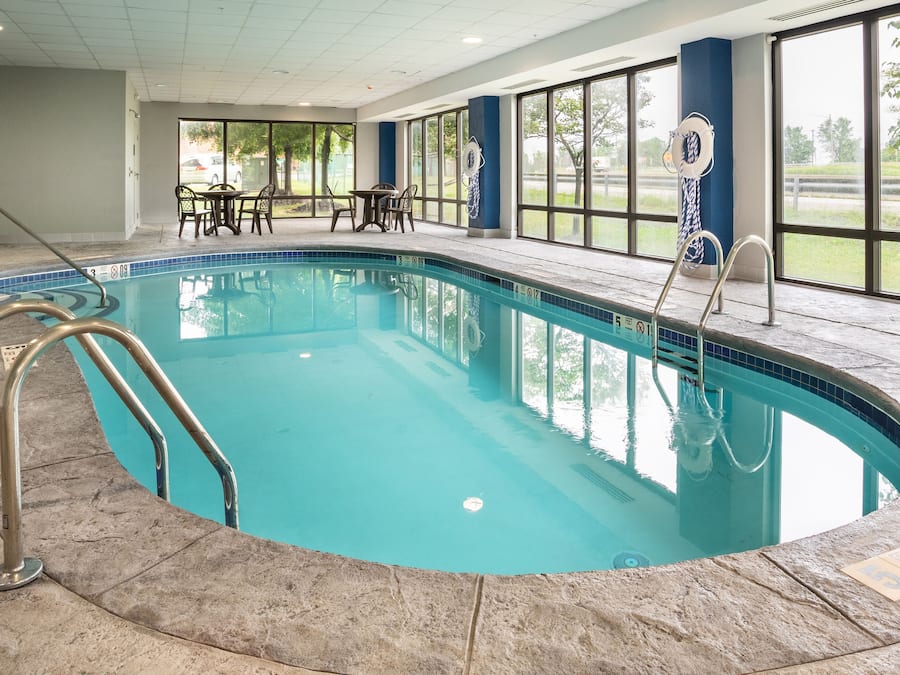 Una piscina interior con agua azul cristalina, rodeada de baldosas de piedra. Se observan escaleras metálicas de piscina, una mesa con sillas y grandes ventanales que ofrecen vistas al exterior.