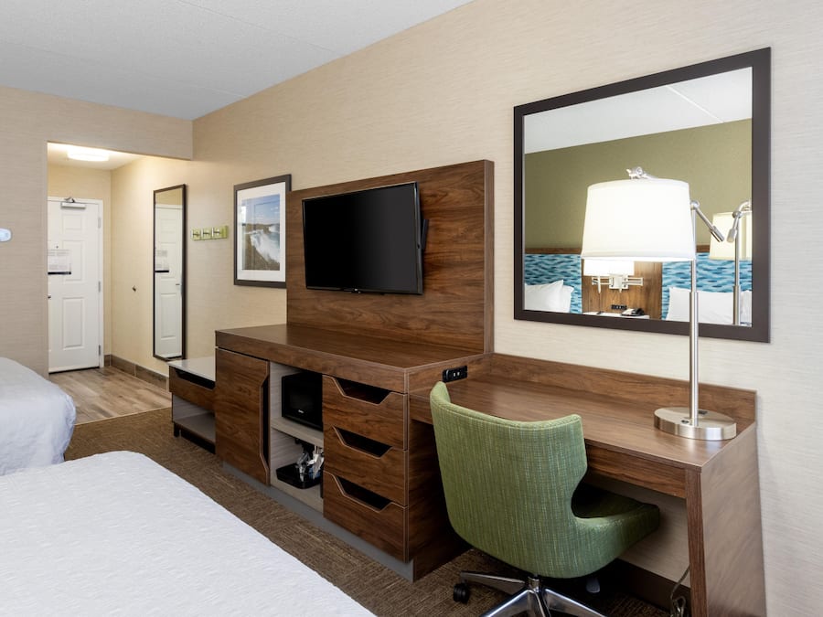 A hotel room with a bed, a desk with a chair, a TV, a mirror, and a lamp.