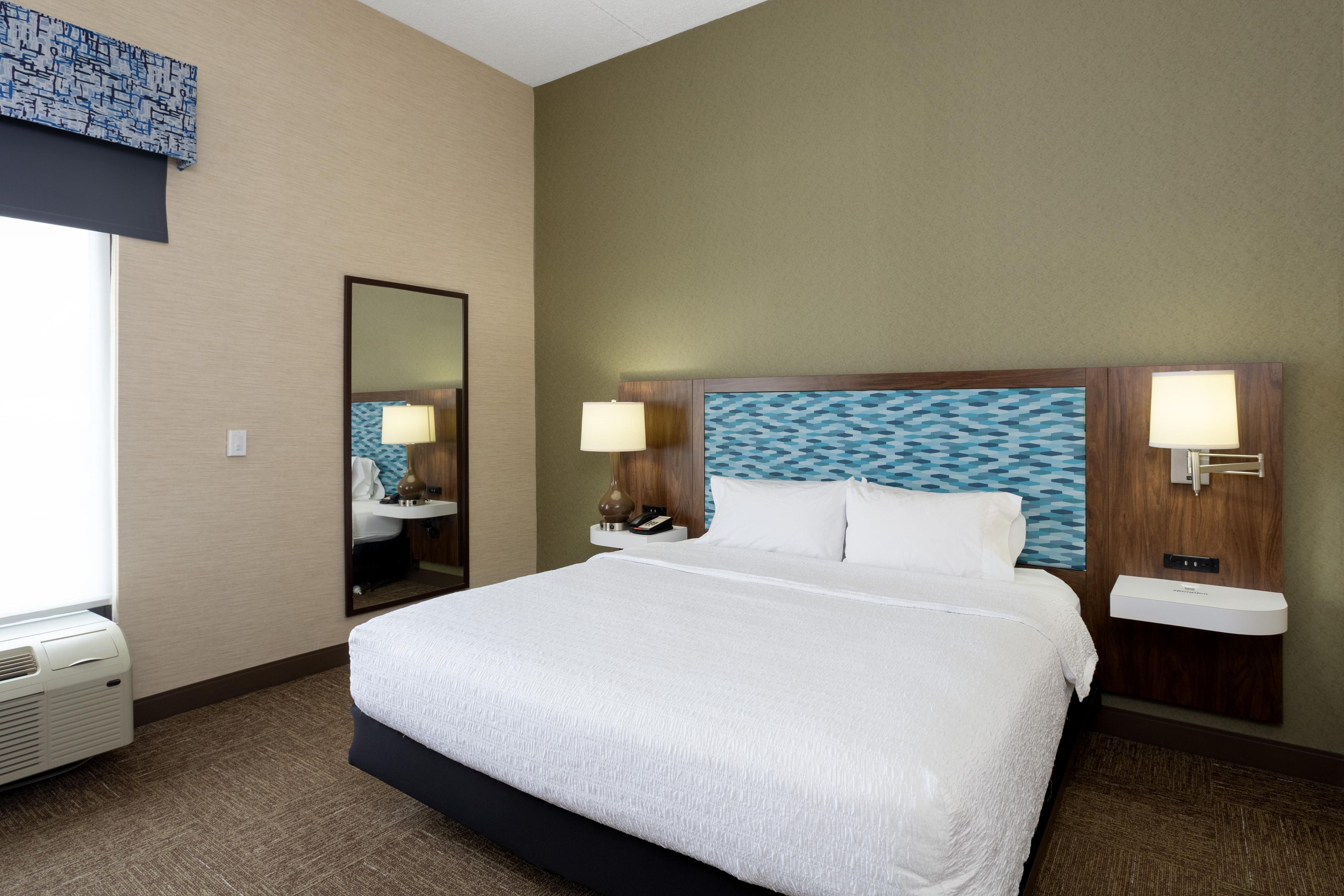 A hotel room with a large bed, two bedside tables, a mirror, a wall-mounted sink, and a window with a view.
