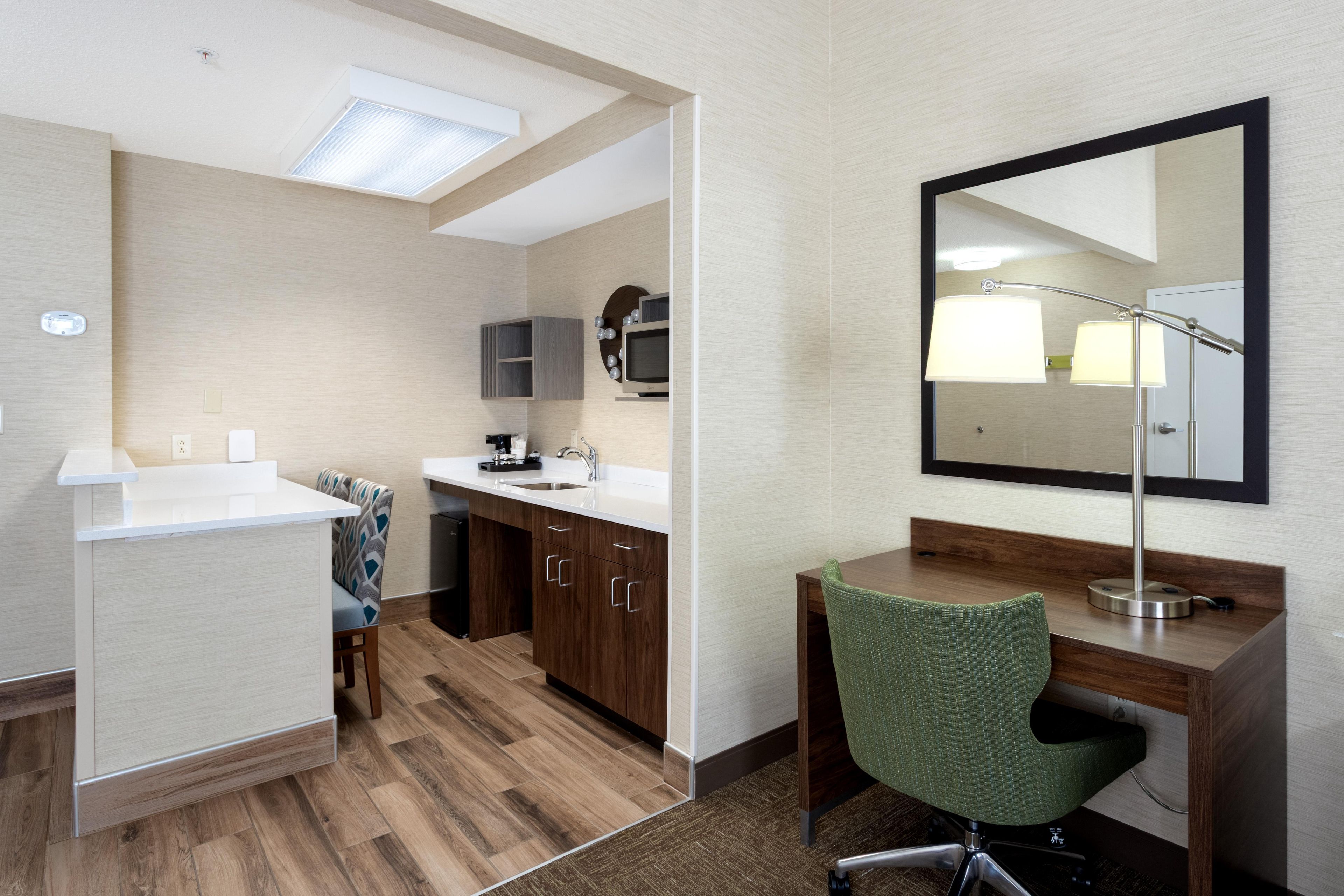 A hotel room with a kitchenette, a desk with a chair, a large mirror, and a ceiling light.