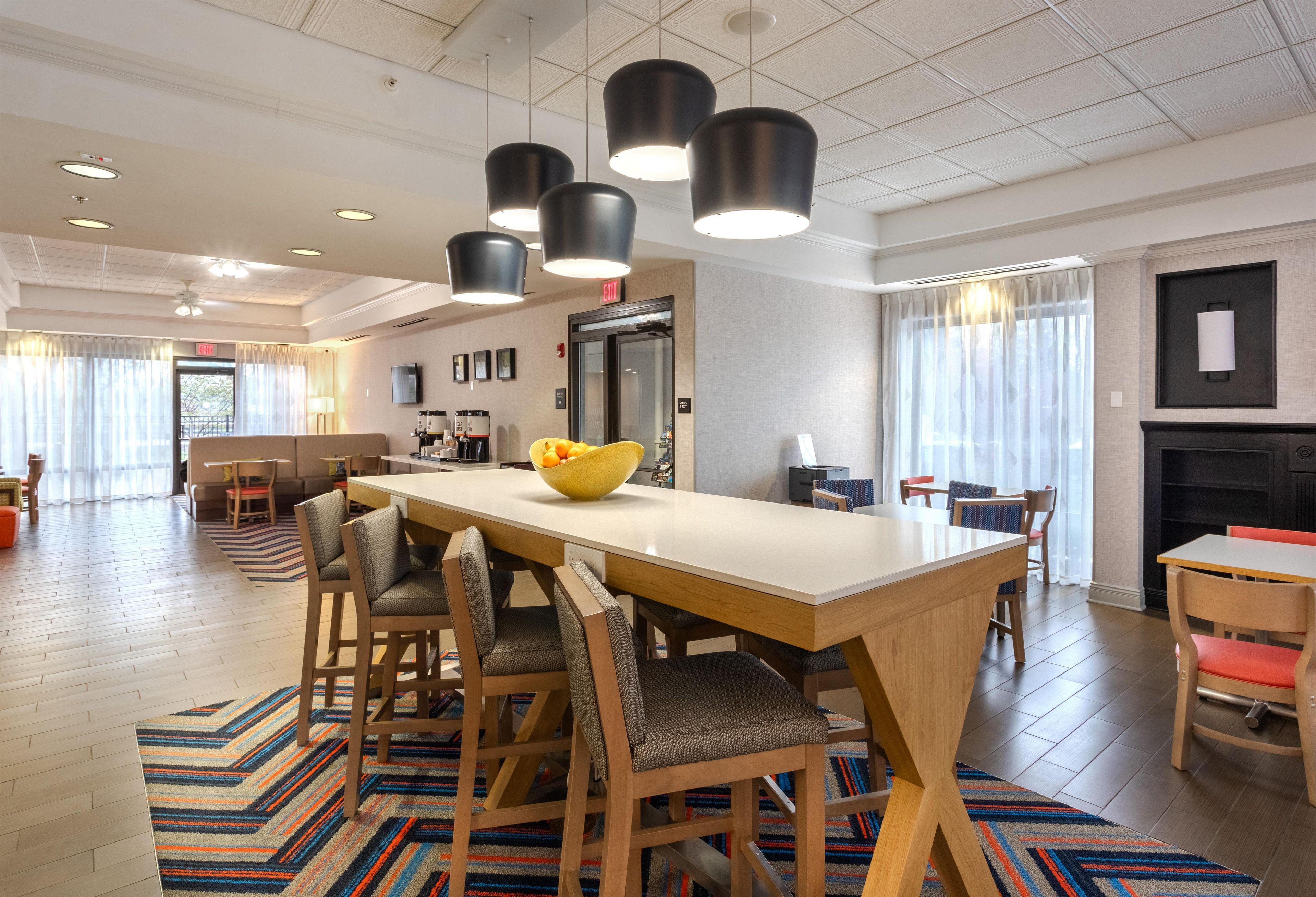 A modern hotel dining area with a large wooden table, multiple chairs, and pendant lighting.